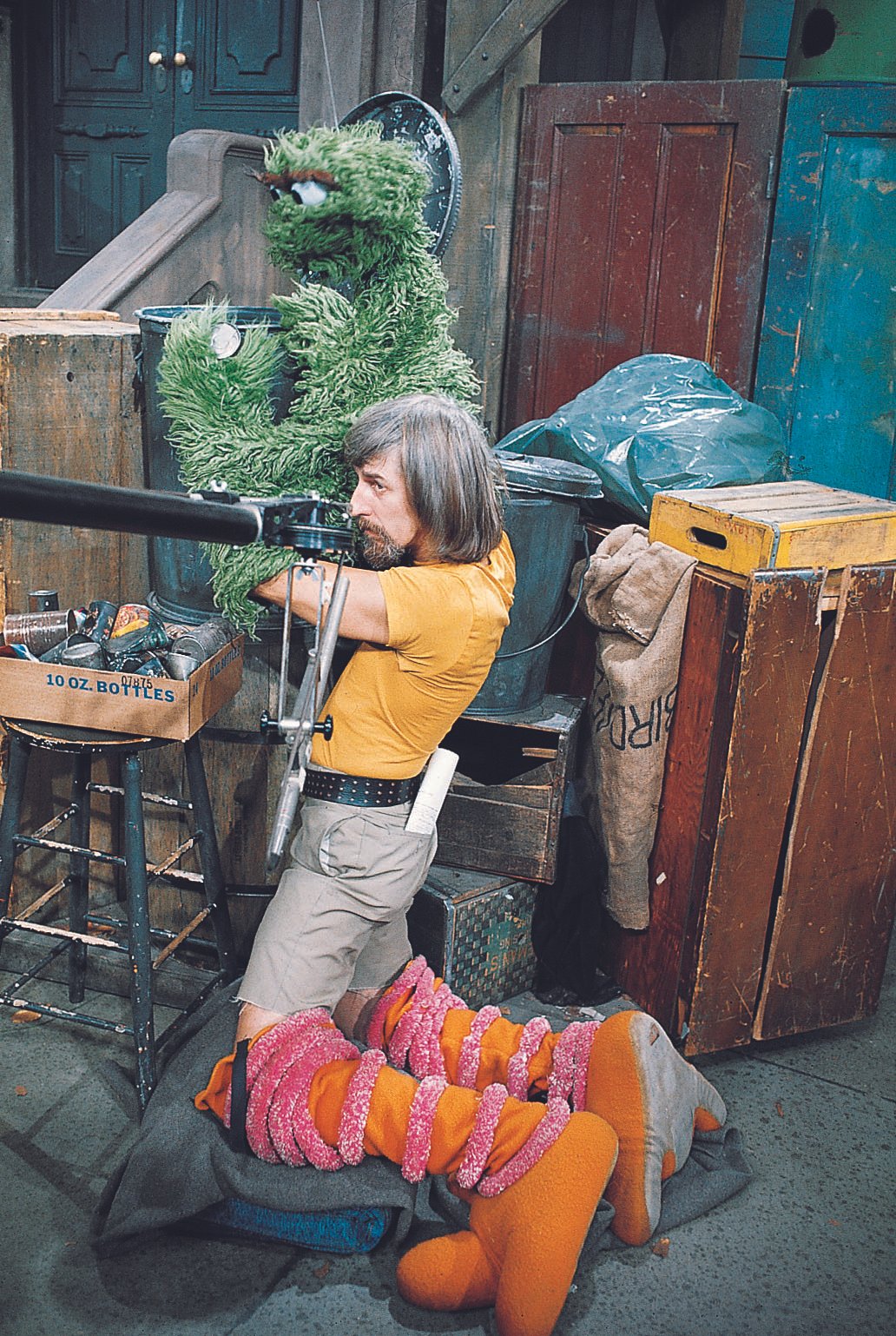 A person dressed in a costume with red, orange, and pink striped pants and large orange shoes is sitting on the floor, leaning against a grey blanket, with their head down. Behind them, a man with long hair and a beard is kneeling and operating a camera on a tripod. To the left, a large green puppet resembling a monster with big eyes and floppy ears is sitting. The scene appears to be behind the scenes of a film or TV production, with wooden crates, bags, and miscellaneous equipment visible around.