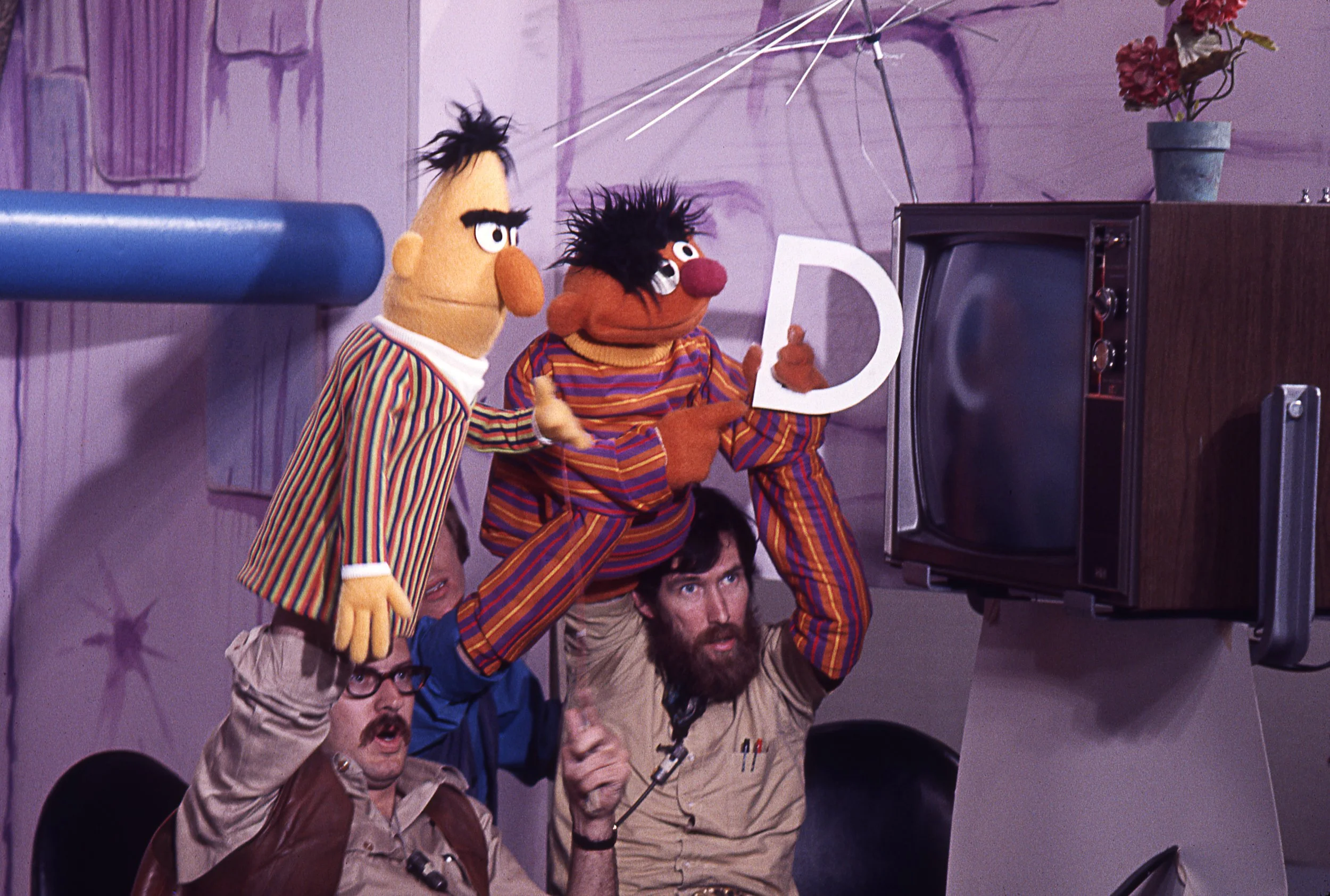 Two Muppets, Bert & Ernie, are being controlled by two men and watching a black-and-white television. Ernie holds a large letter D, and the background is purple with stars painted on the wall.