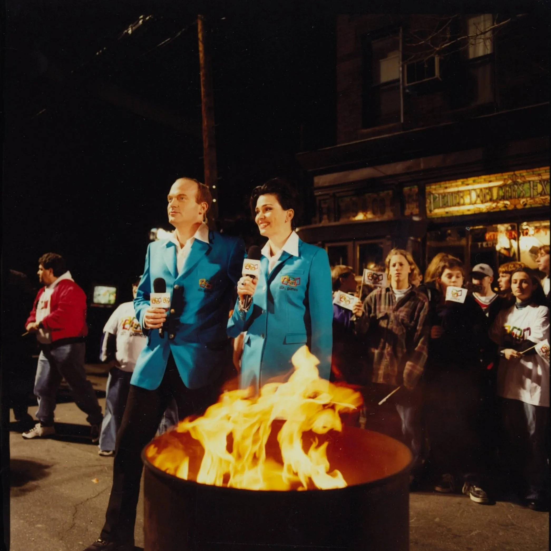 Tad Low & Karen Duffy in blue suits standing in front of a crowd during a nighttime outdoor event, with a fiery bonfire in the foreground, holding microphones with logo.