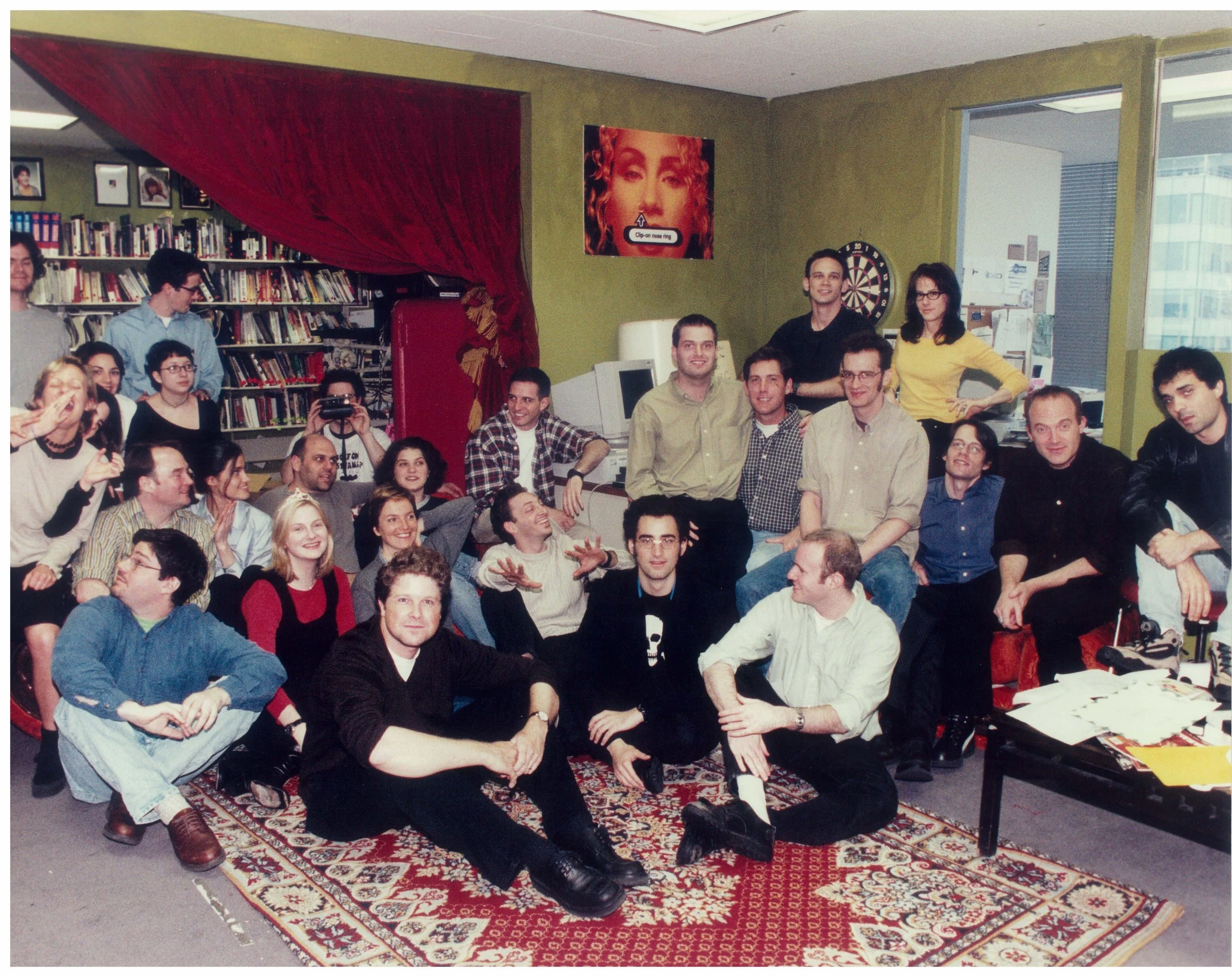 The staff of "Pop-Up Video" gathered in an office space, sitting on and around a patterned rug, with a bookshelf, computer, and dartboard in the background, and a window showing city buildings.