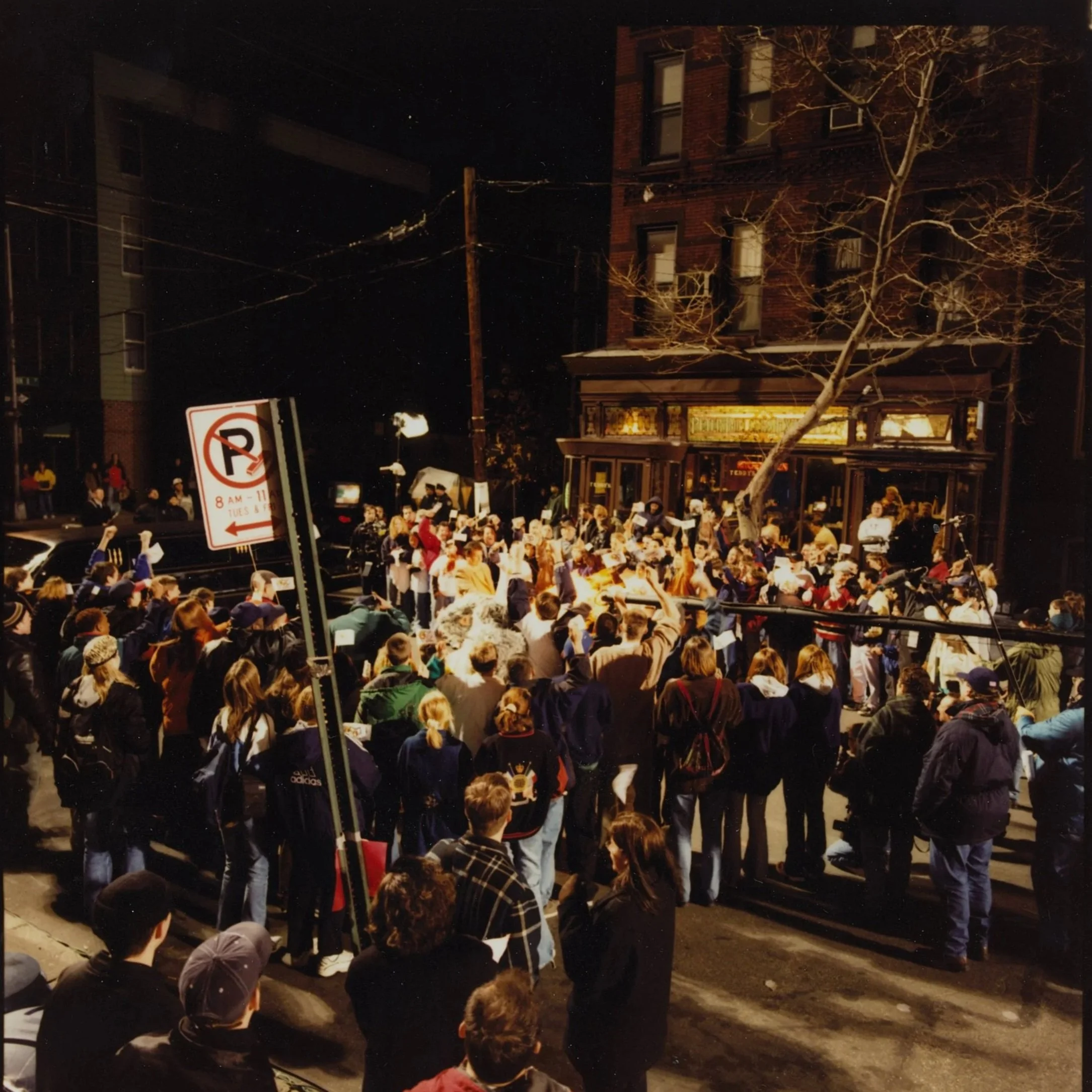 Crowd gathered outside a restaurant at night, some people holding umbrellas, with street parking signs and buildings visible in the background.