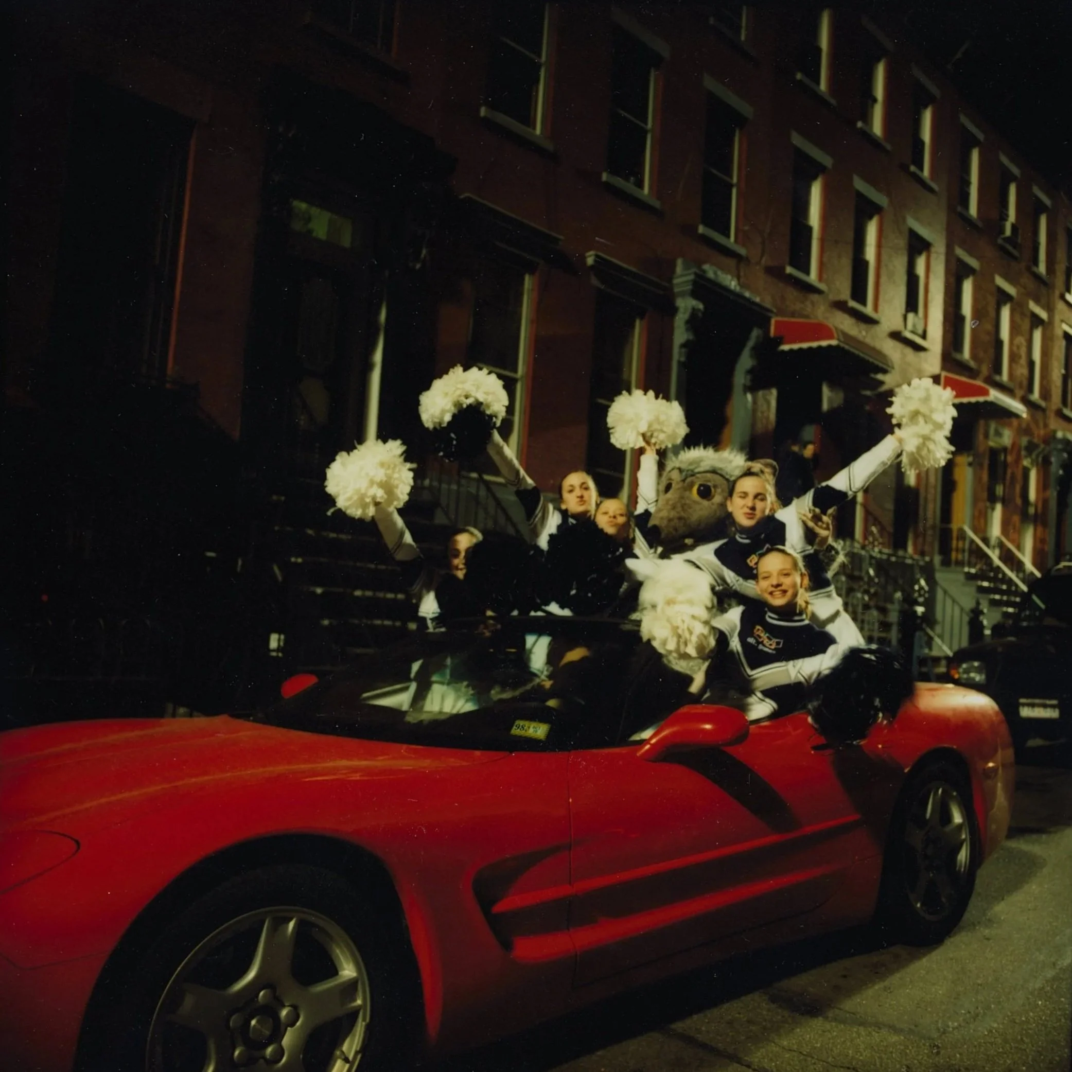 Cheerleaders riding in a red convertible car decorated for a parade at night, with a large rat mascot and cheer pom-poms, in front of brownstone buildings.
