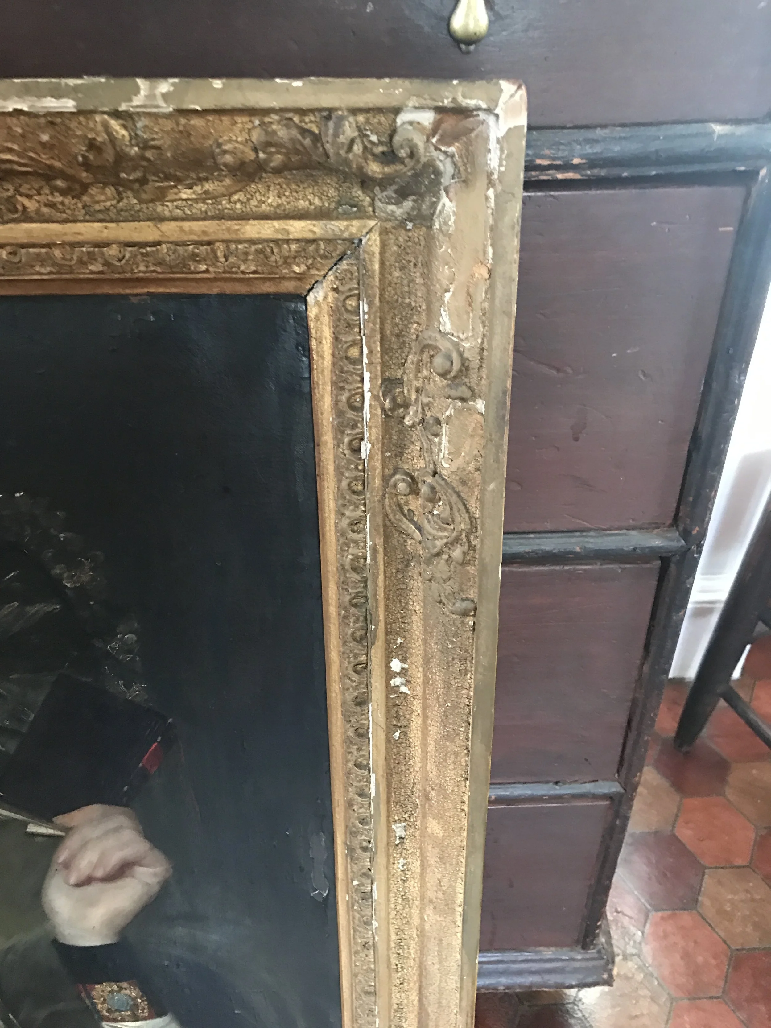 Close-up of an ornate, gold-colored picture frame corner with intricate detailing, next to a dark wooden cabinet, and part of a person’s hand holding a black object in the bottom left corner.