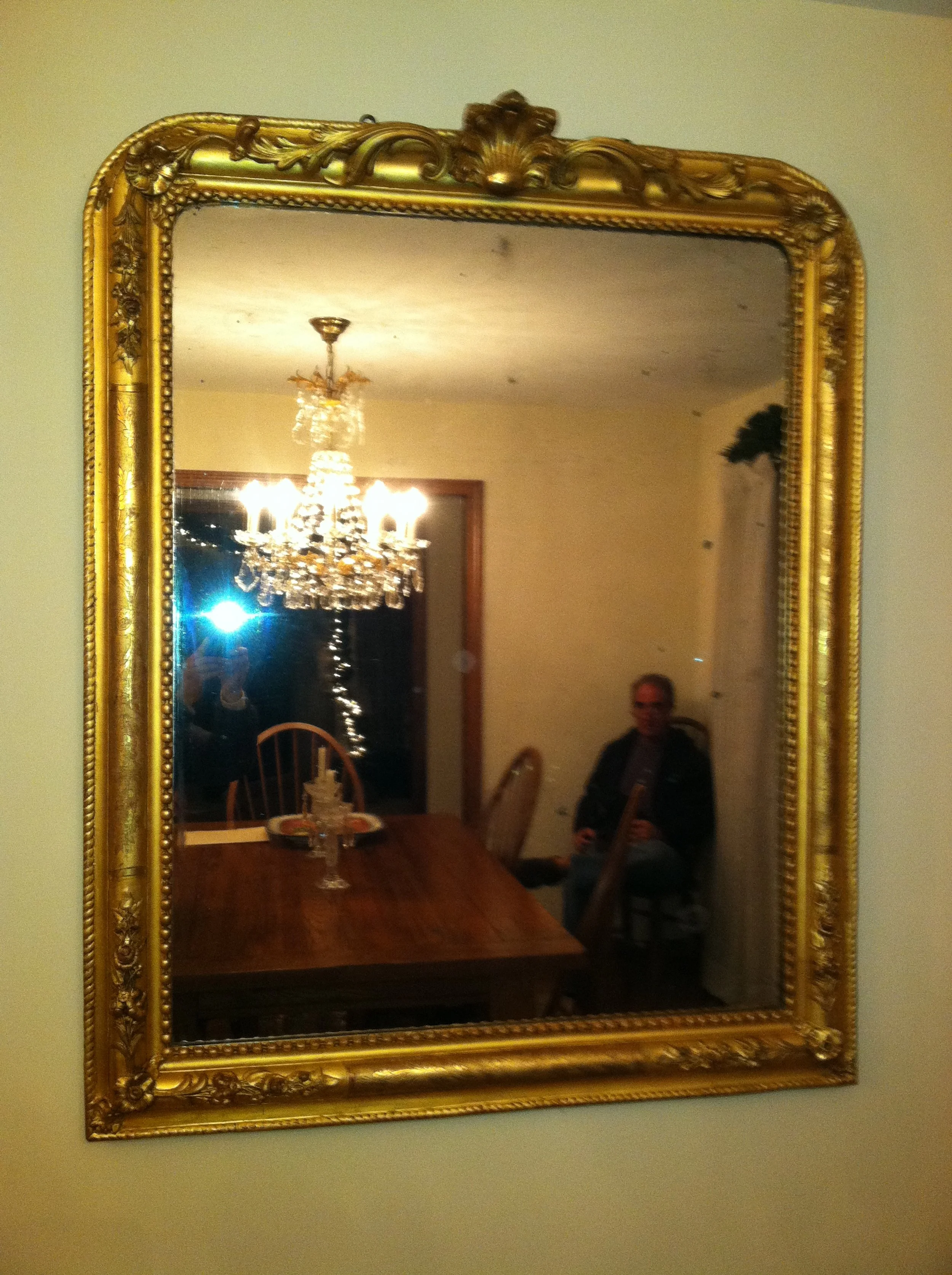 A gold ornate mirror reflecting a chandelier hanging over a wooden dining table with chairs, a person seated in a corner, and a window with curtains.