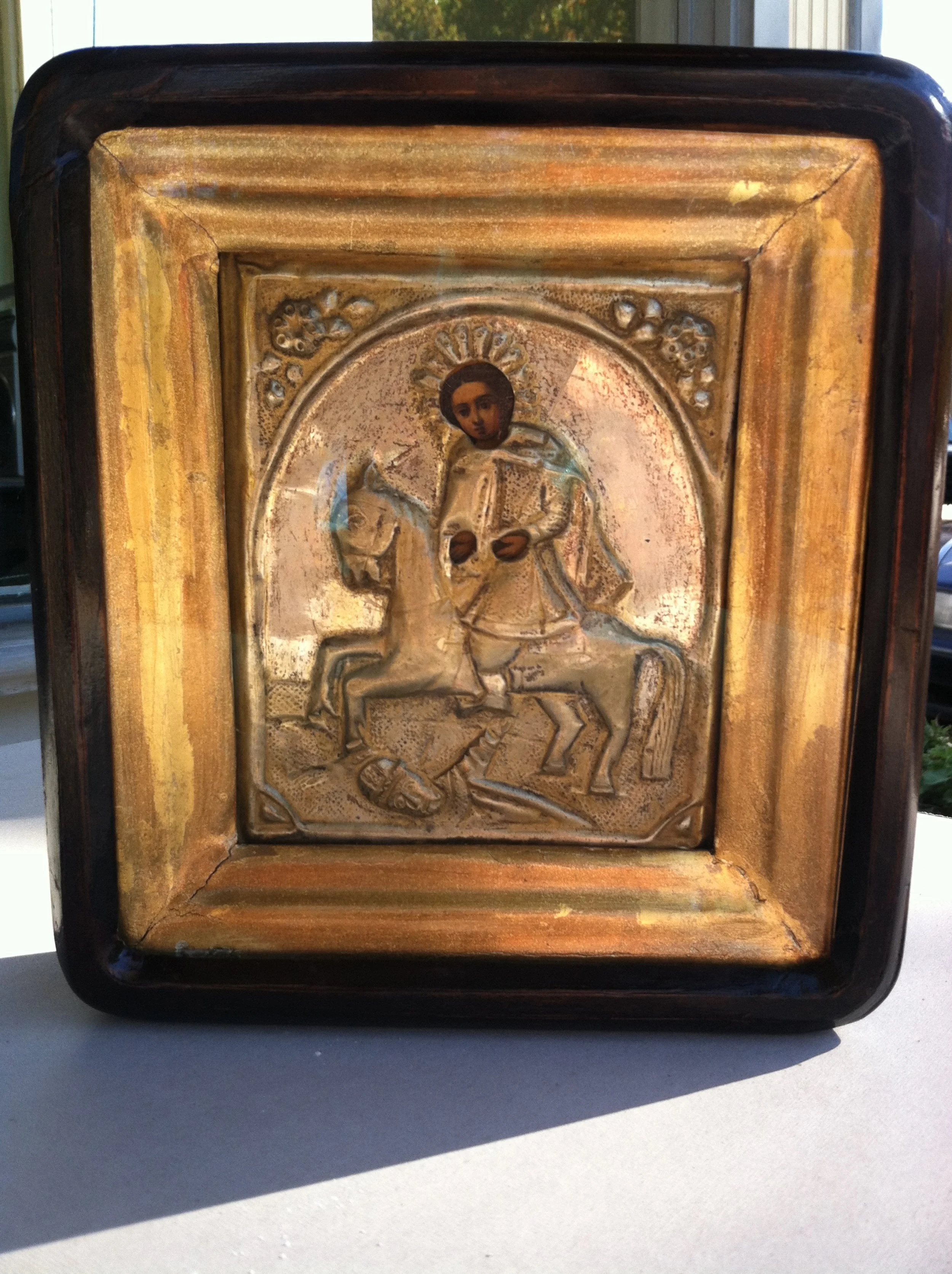 A religious icon depicting a saint on a horse, holding a sword and shield, with a halo around his head, set in a decorative metal frame with a wooden border.