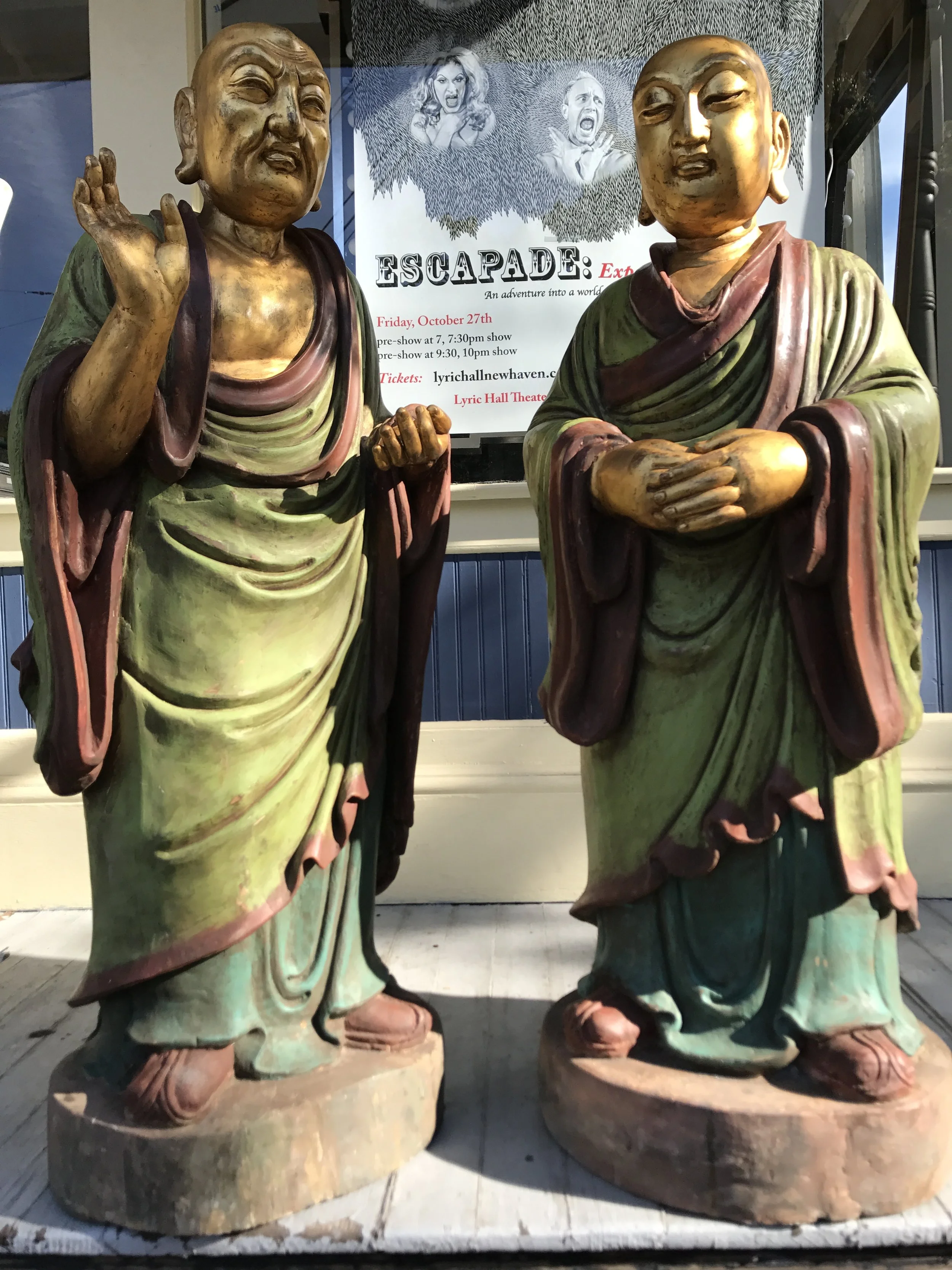 Two life-sized statues of Buddhist monks, one raising his hand and the other with hands clasped, with a poster about an event titled 'ESCAPADE' in the background.