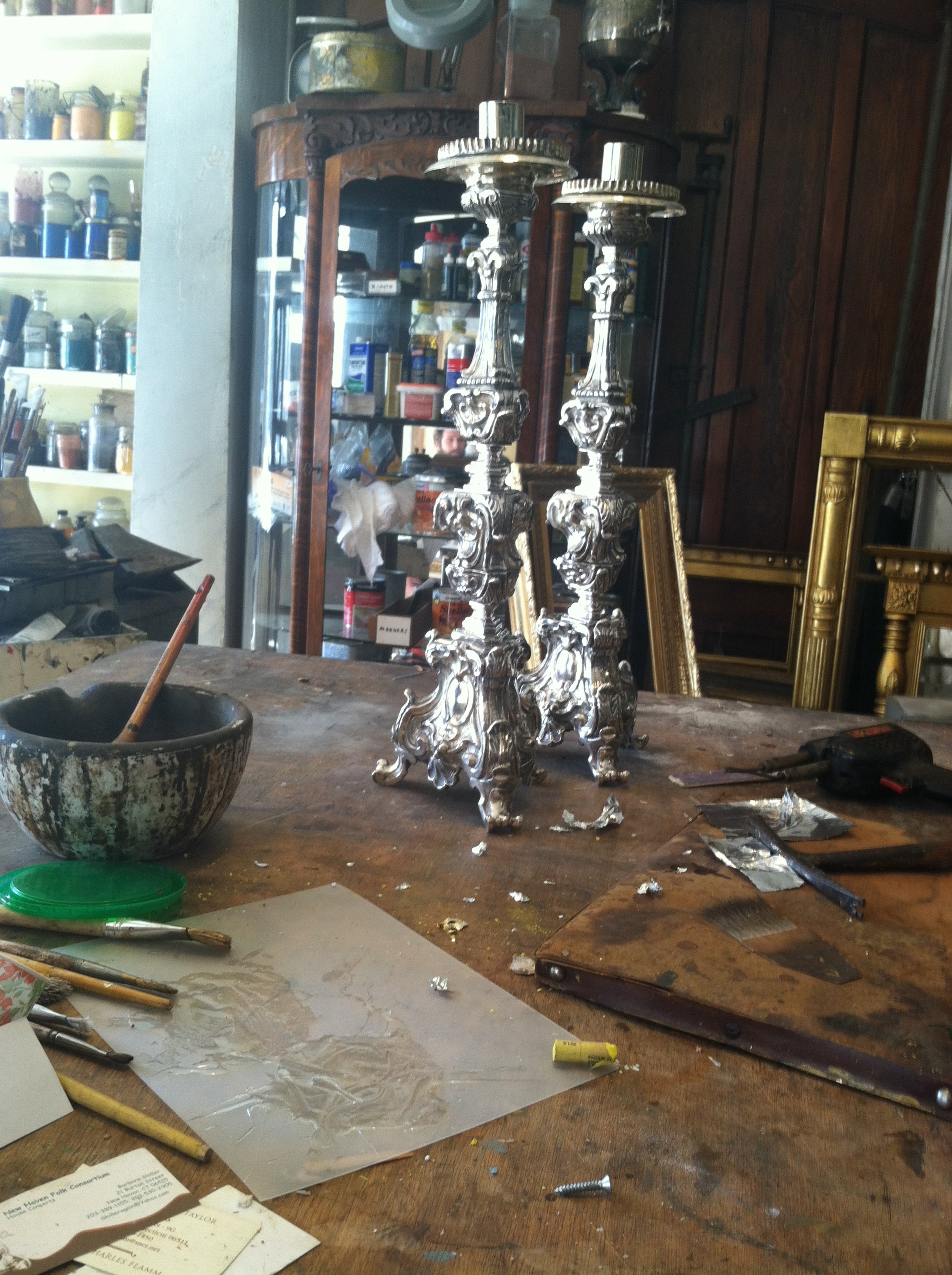 A cluttered worktable with silver ornate candlesticks, a bowl with tools, and various art supplies in a workshop.