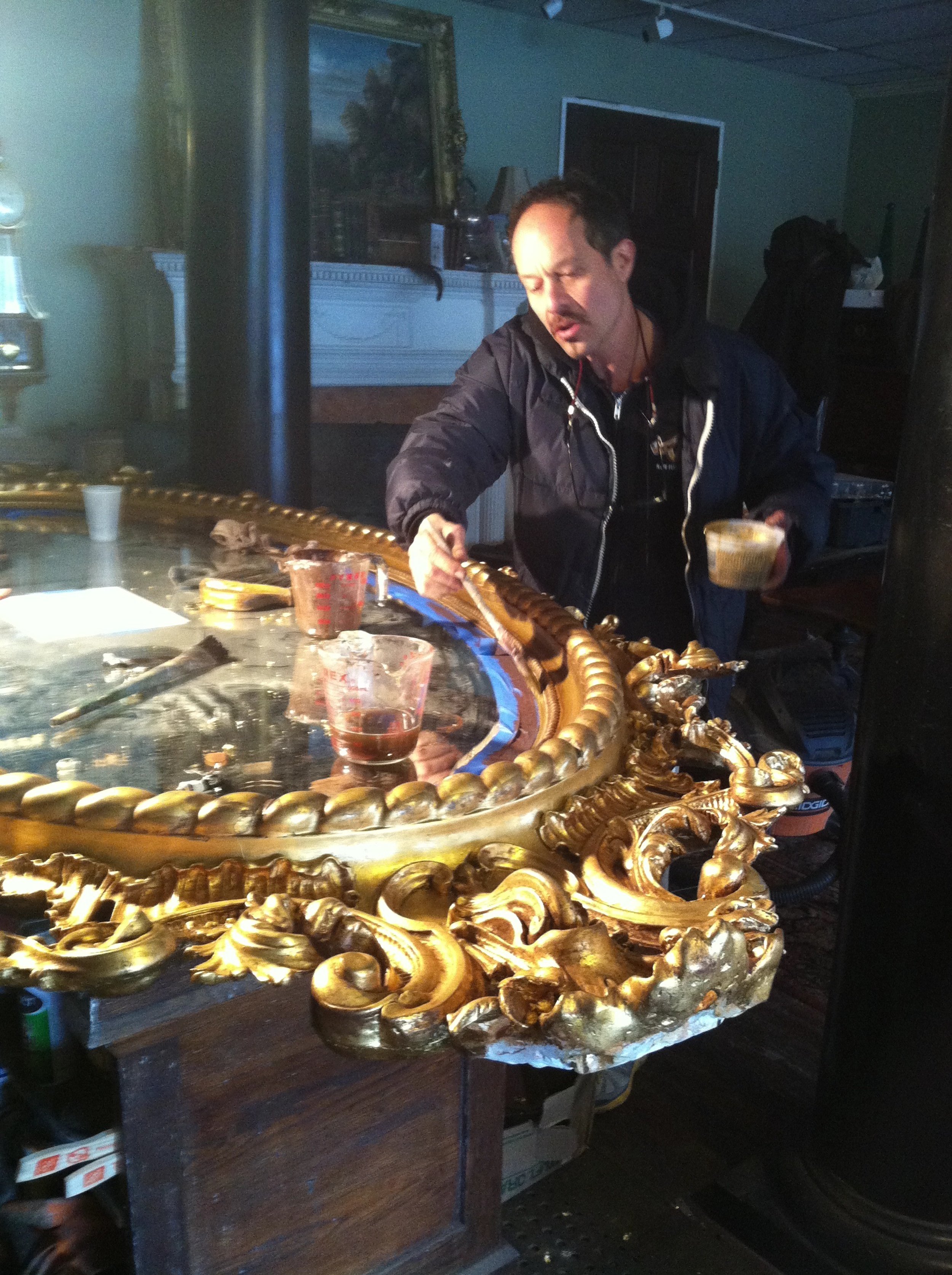 A man wears a dark jacket and is cleaning a golden, ornate mirror frame with a brush, standing behind a decorated table with glue, cups, and tools, in a room with paintings, a white fireplace, and a TV.