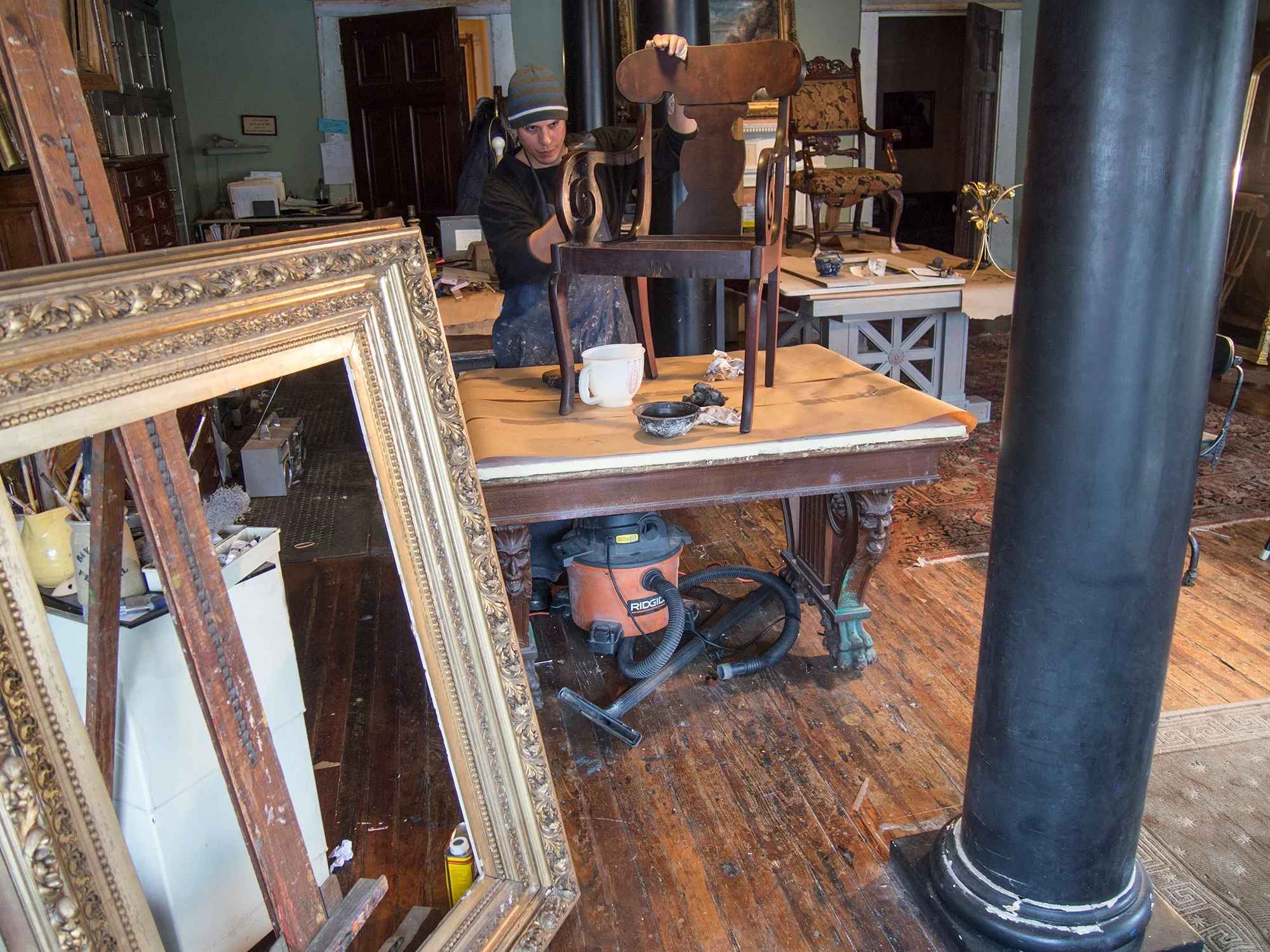 A person working in an antique furniture restoration workshop, surrounded by various furniture pieces, tools, and decorative items.