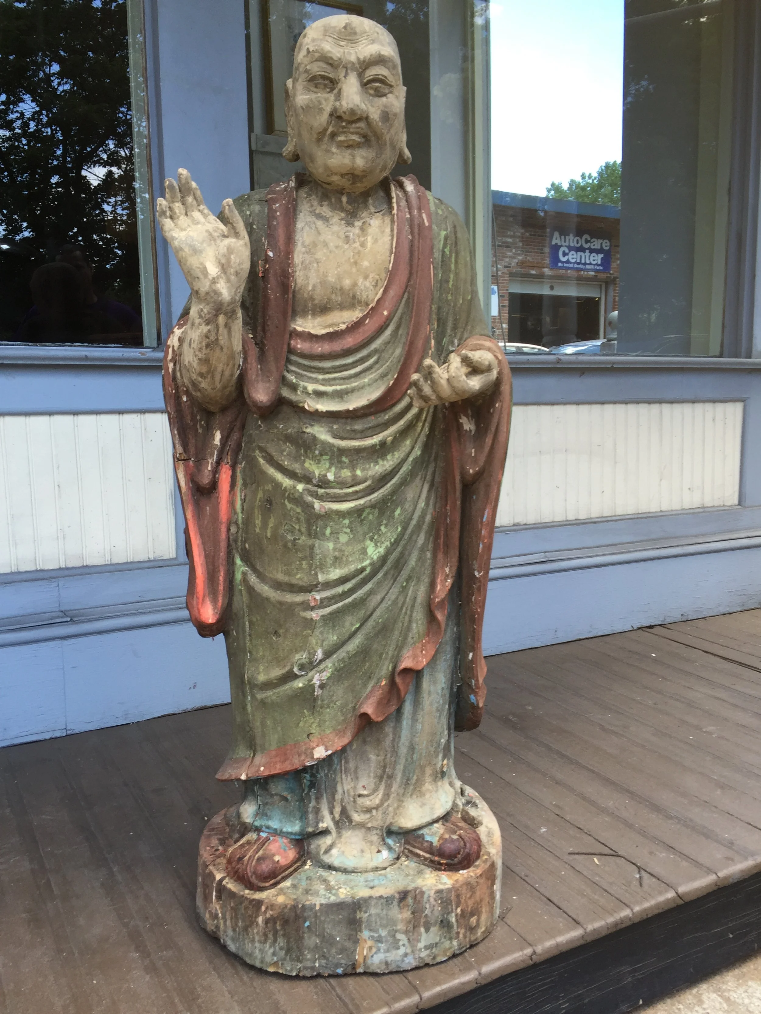 Weathered wooden statue of a standing man in traditional robe, raising his right hand in greeting or blessing, placed on a wooden surface outside a building.