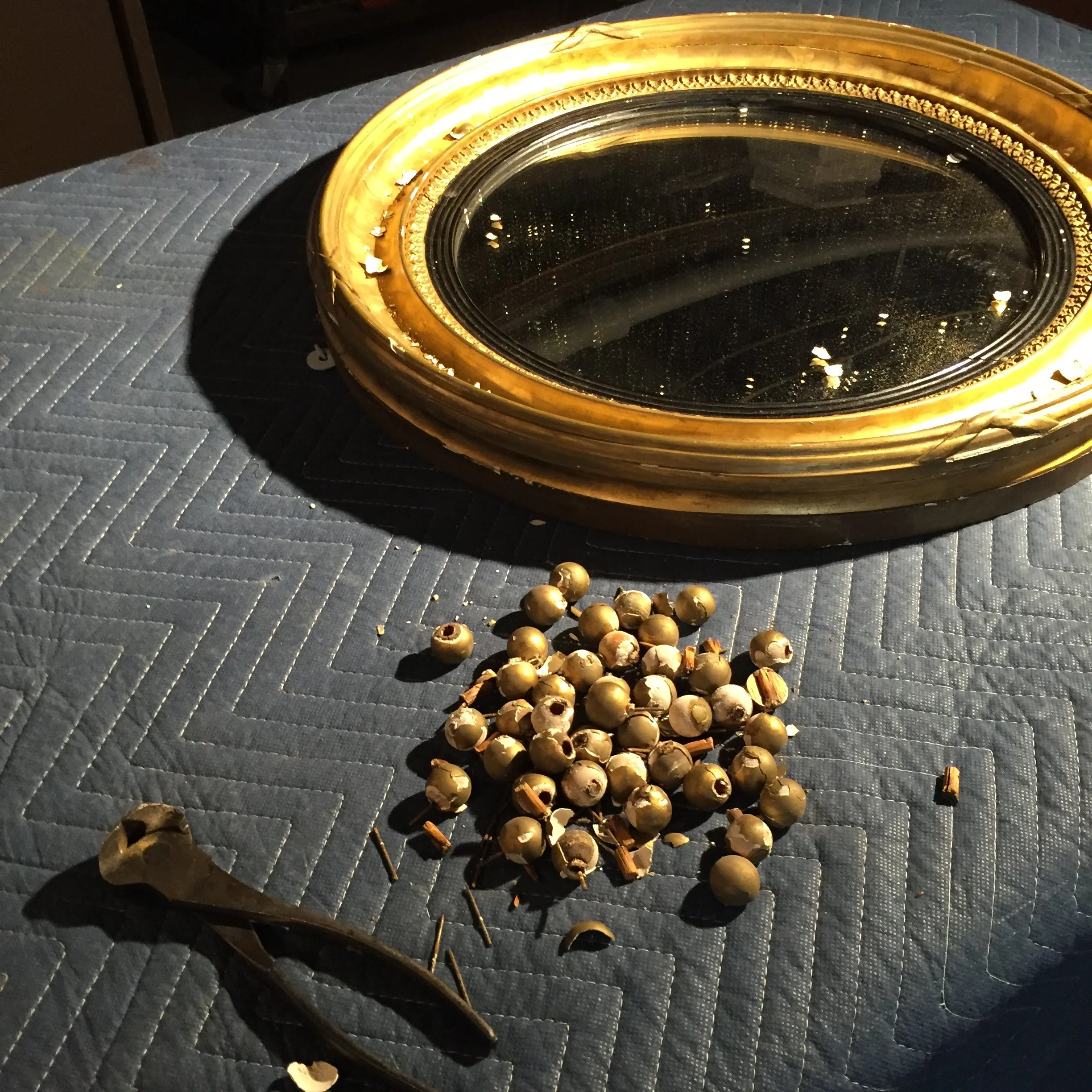 A large, ornate, oval mirror with a gold frame placed on a textured dark blue quilted surface. In front of the mirror, there is a small pile of dried berries and a wooden spoon.