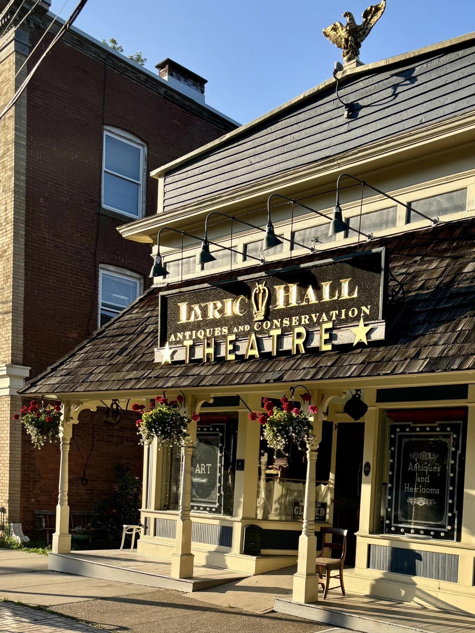 Front view of the vintage-style storefront for Lyric Hall Restoration, Antiques and Conservation Theatre, with hanging flower baskets and an illuminated sign.