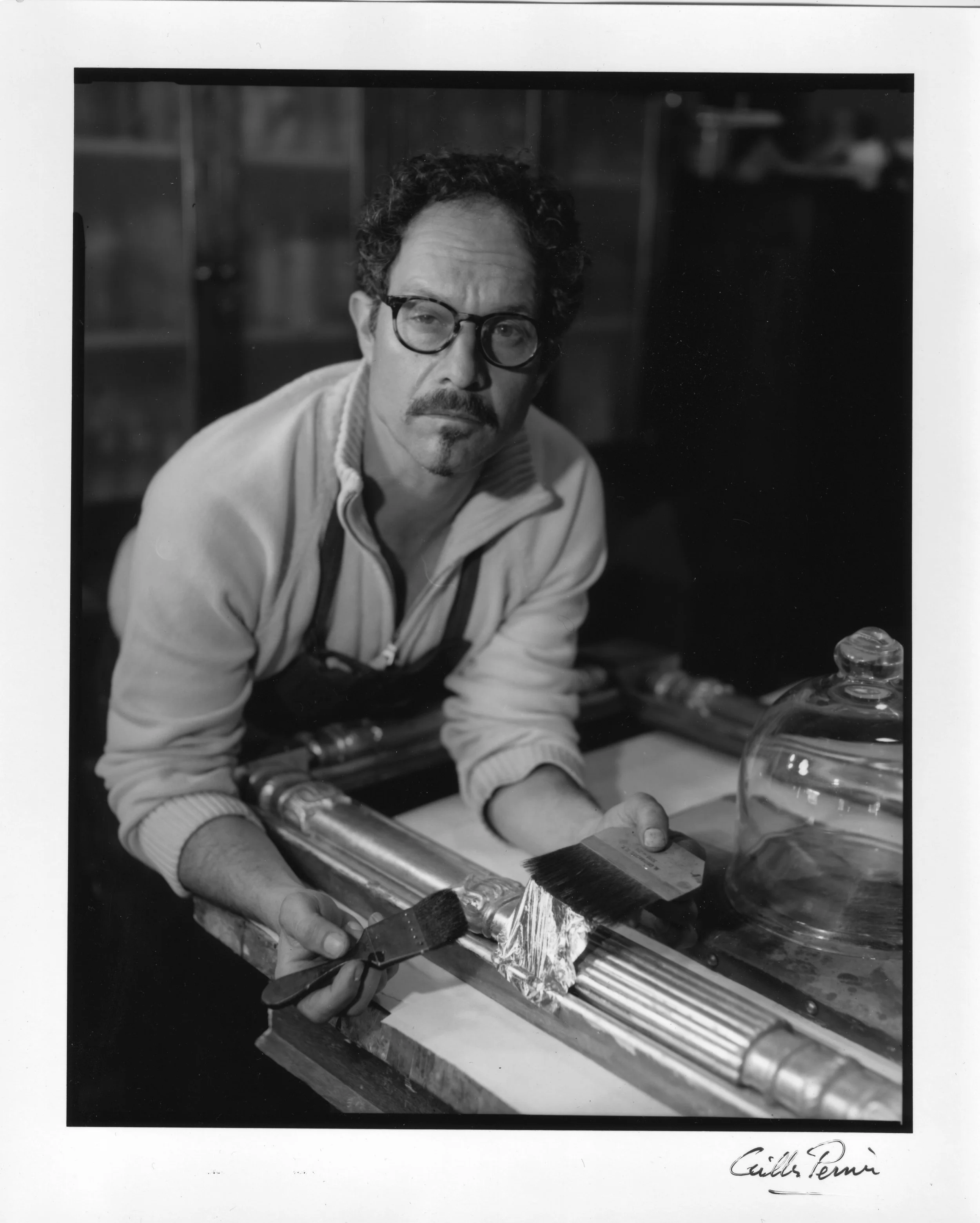 Black and white photo of a man with curly hair, glasses, and a mustache working on a glass art piece in a workshop. He is holding a brush and appears to be applying or polishing the glass frame.