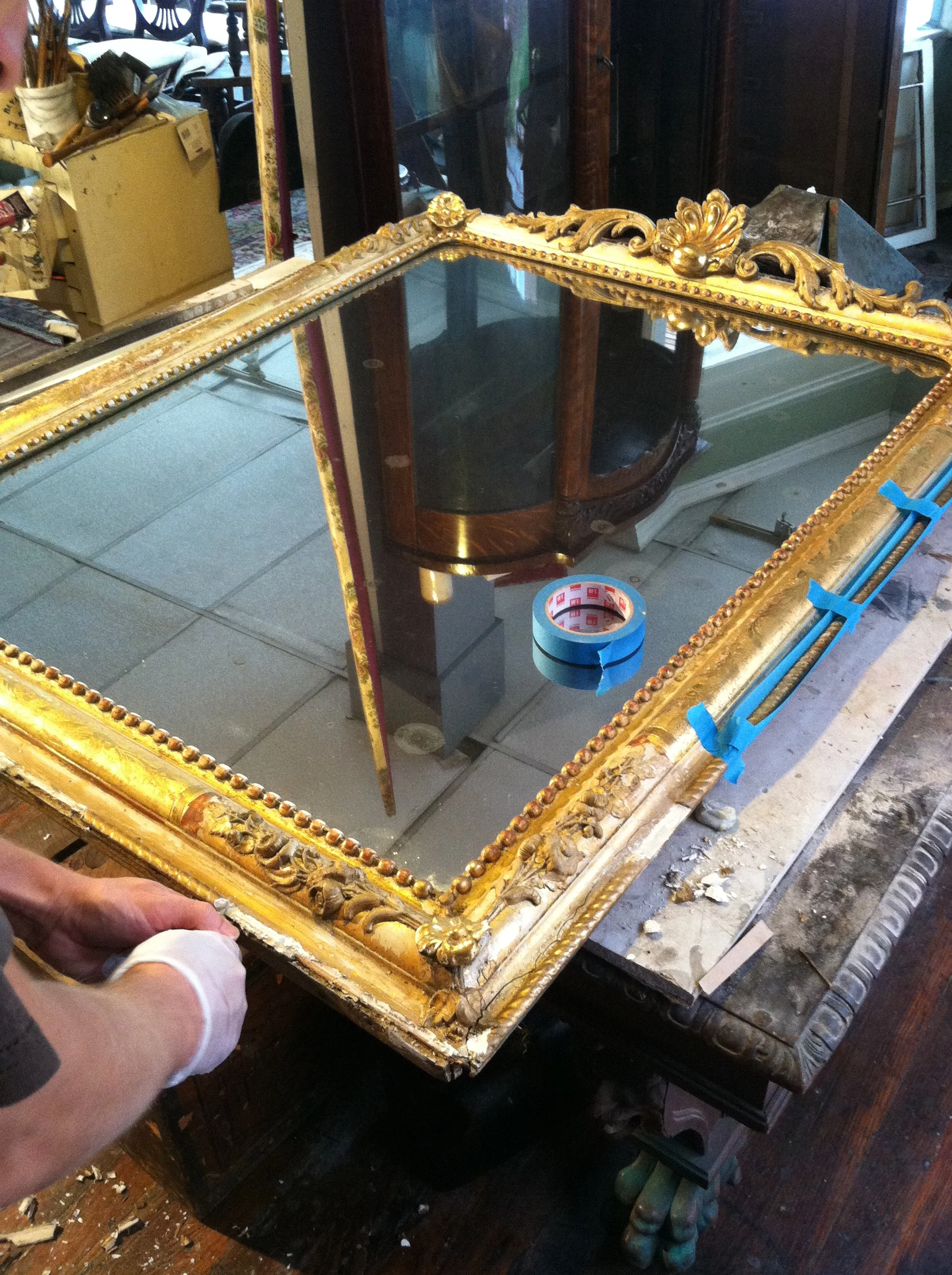 A person working on restoring a vintage gold-framed mirror with ornate carvings, placed on a wooden table, in a cluttered workshop.