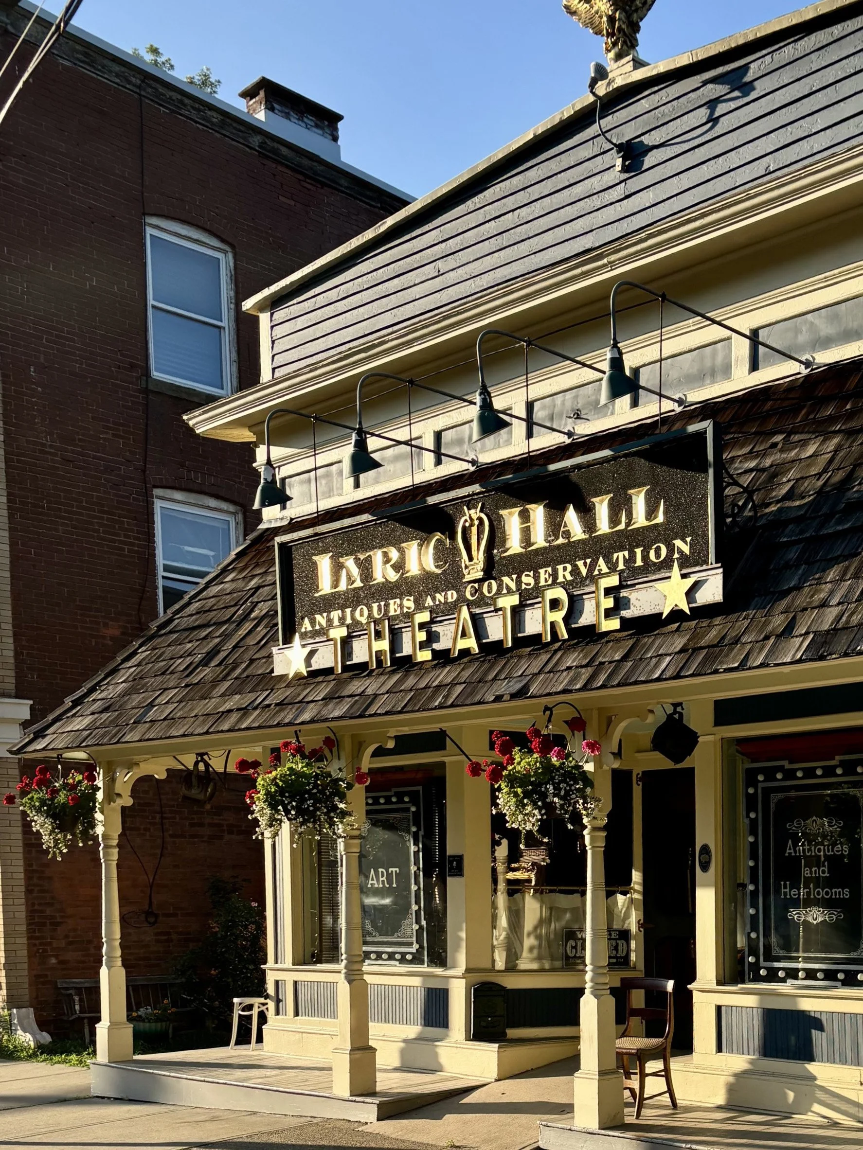 Facade of Lyric Hall, an antique and conservation theater, with hanging flower baskets, a porch, and signage indicating antiques and heirlooms.