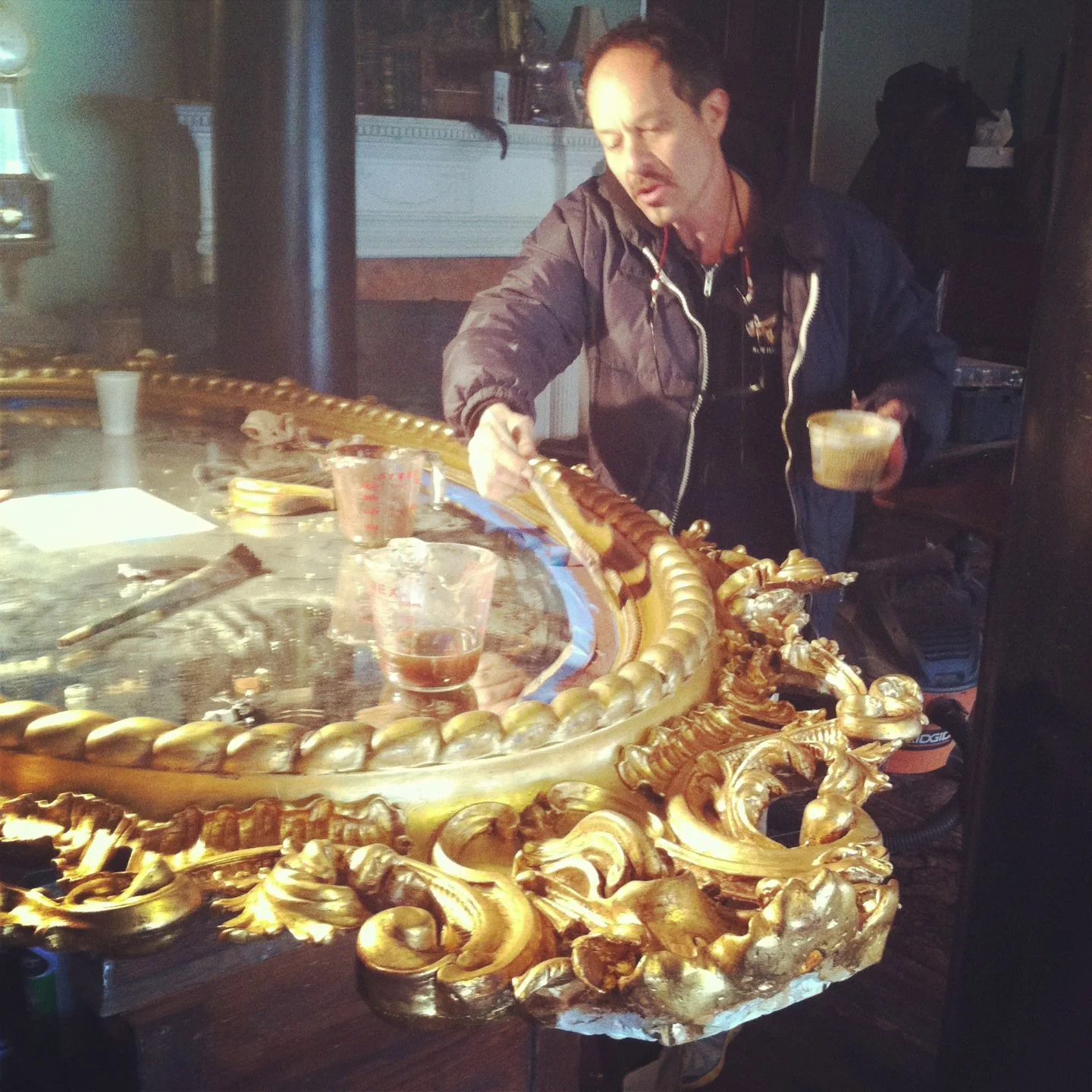 A man working on an ornate, gold-painted, carved frame or decoration near a glass-topped table with drinks and tools.