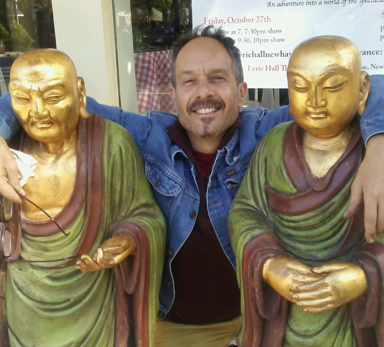 A man with a beard and mustache, wearing a denim jacket, smiling and sitting between two statues of monks with gold faces and robes, outside a building with a sign in the background.
