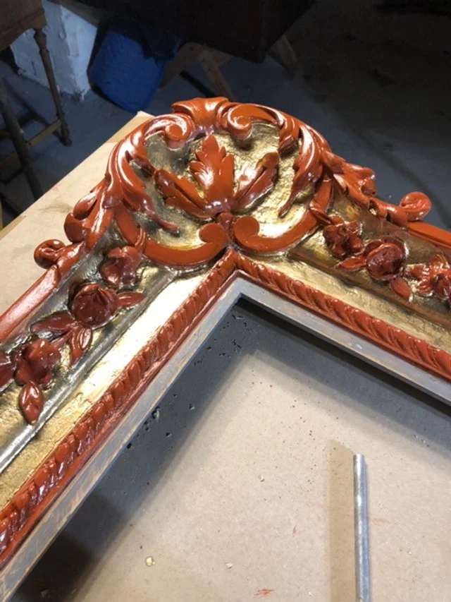Close-up of an ornate, carved wooden frame with gold and reddish-brown painted details, lying on a worktable.