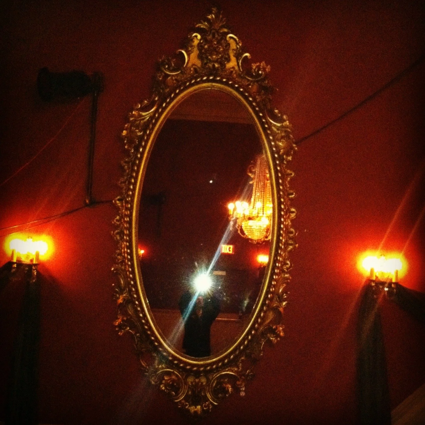 Ornate gold-framed oval mirror on a red wall, reflecting part of a chandelier and the photographer taking the picture with a flash.