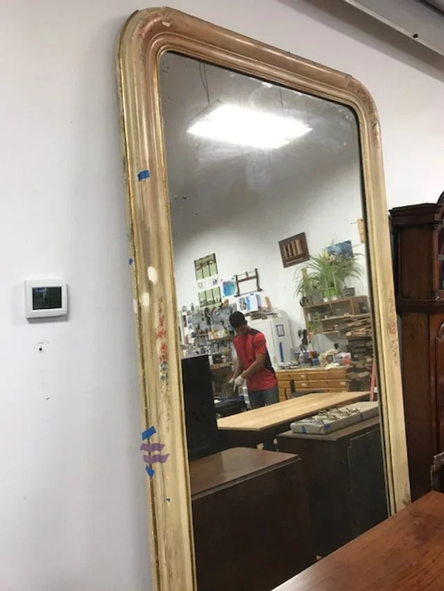 A large vintage mirror with a yellowed frame leaning against a white wall, reflecting a store interior with a person in a red shirt browsing.
