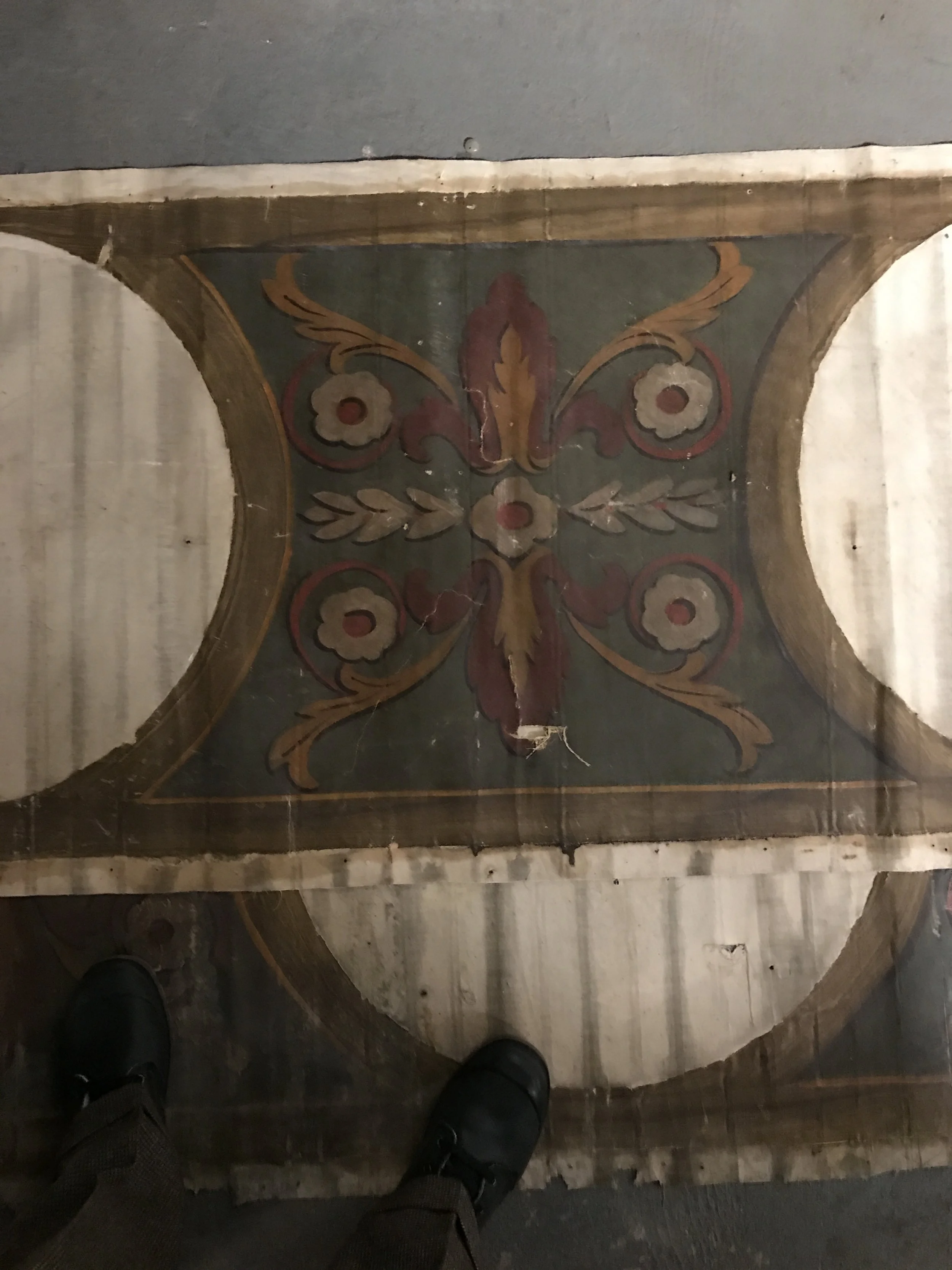 Vintage decorative tiled floor with floral and scroll design, showing part of a person's shoes and pants.