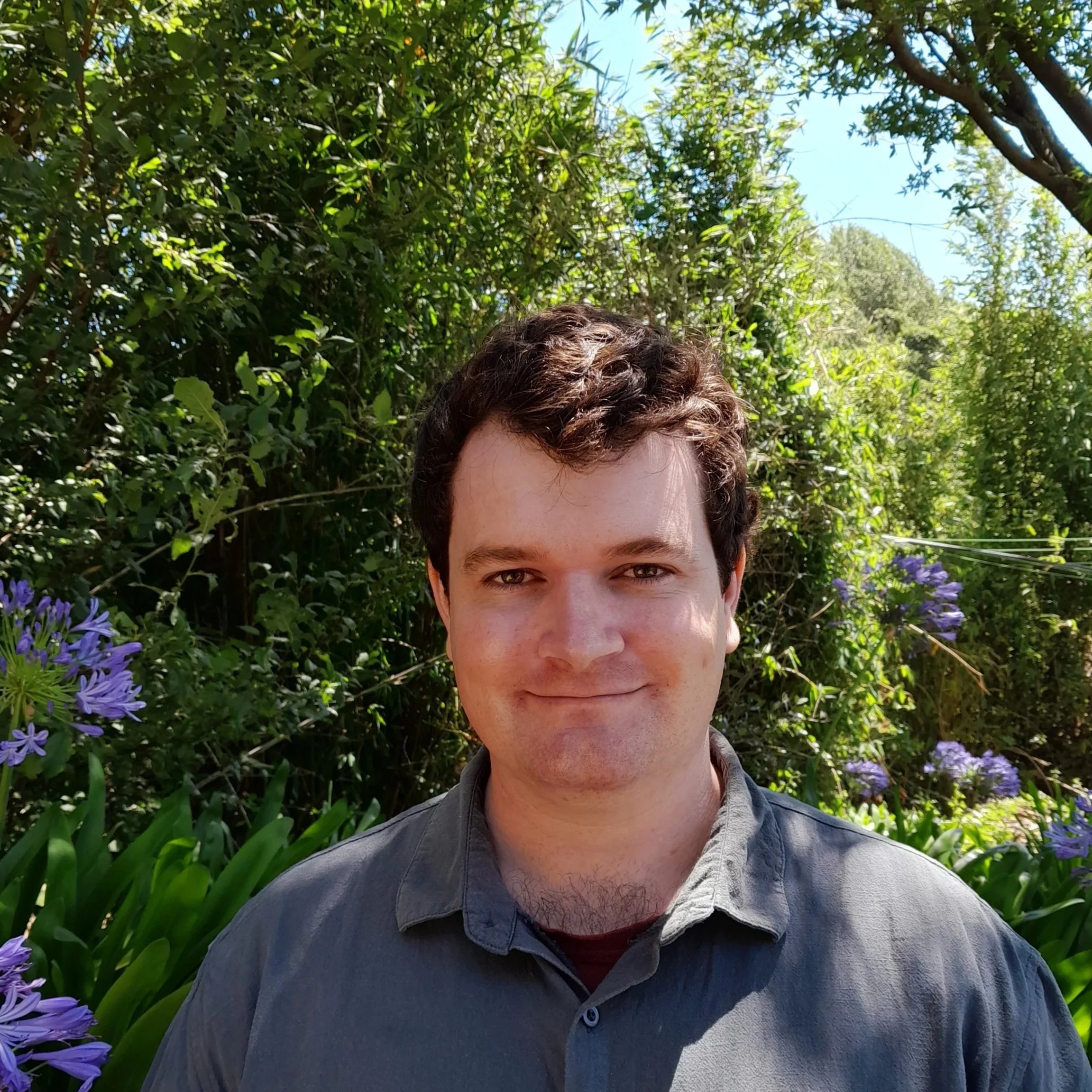 A young man with curly brown hair and light skin, smiling, standing outdoors in a lush green garden with purple flowers and trees in the background on a sunny day.