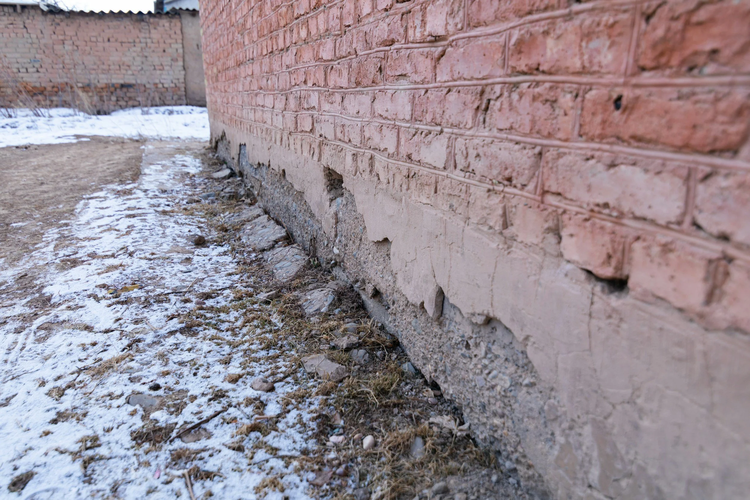 Close-up of a damaged brick wall with crumbling concrete at the base, and patches of snow and dirt on the ground outside.