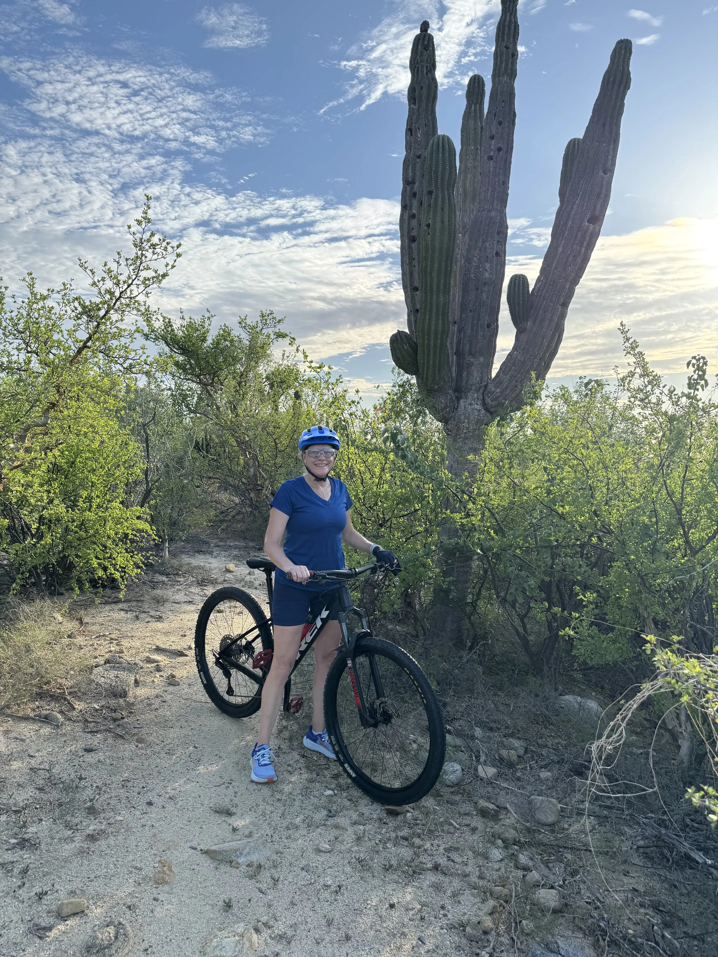 A woman in a blue shirt, shorts, and a helmet standing with a mountain bike on a dirt trail in a desert landscape with a large saguaro cactus and green bushes, under a partly cloudy sky during daytime.