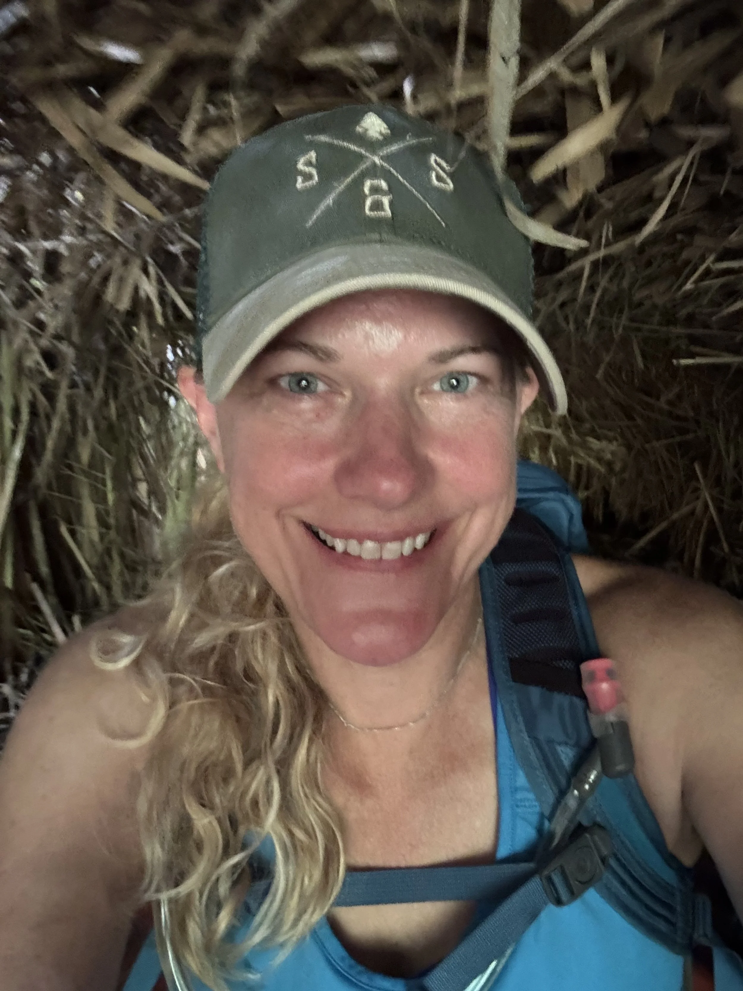 A woman with curly blonde hair smiling while taking a selfie in a natural outdoor setting surrounded by branches and foliage, wearing a grey cap and a blue tank top with a backpack.