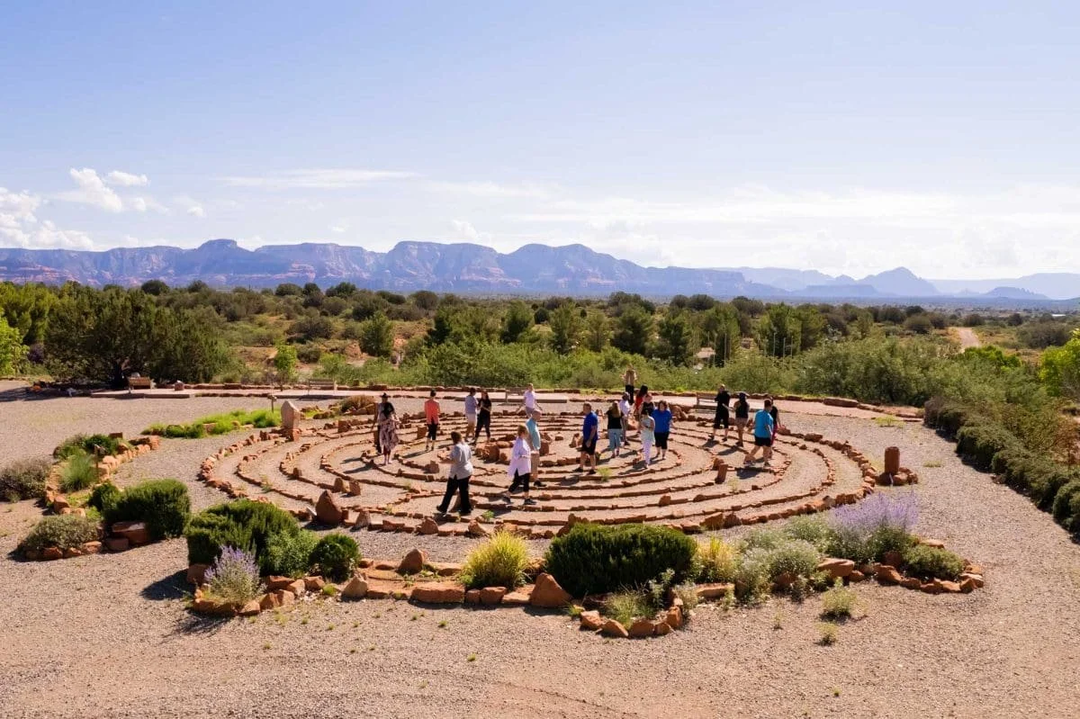 People walking in a labyrinth at Sedona Mago made of stones in a desert landscape with mountains in the background.