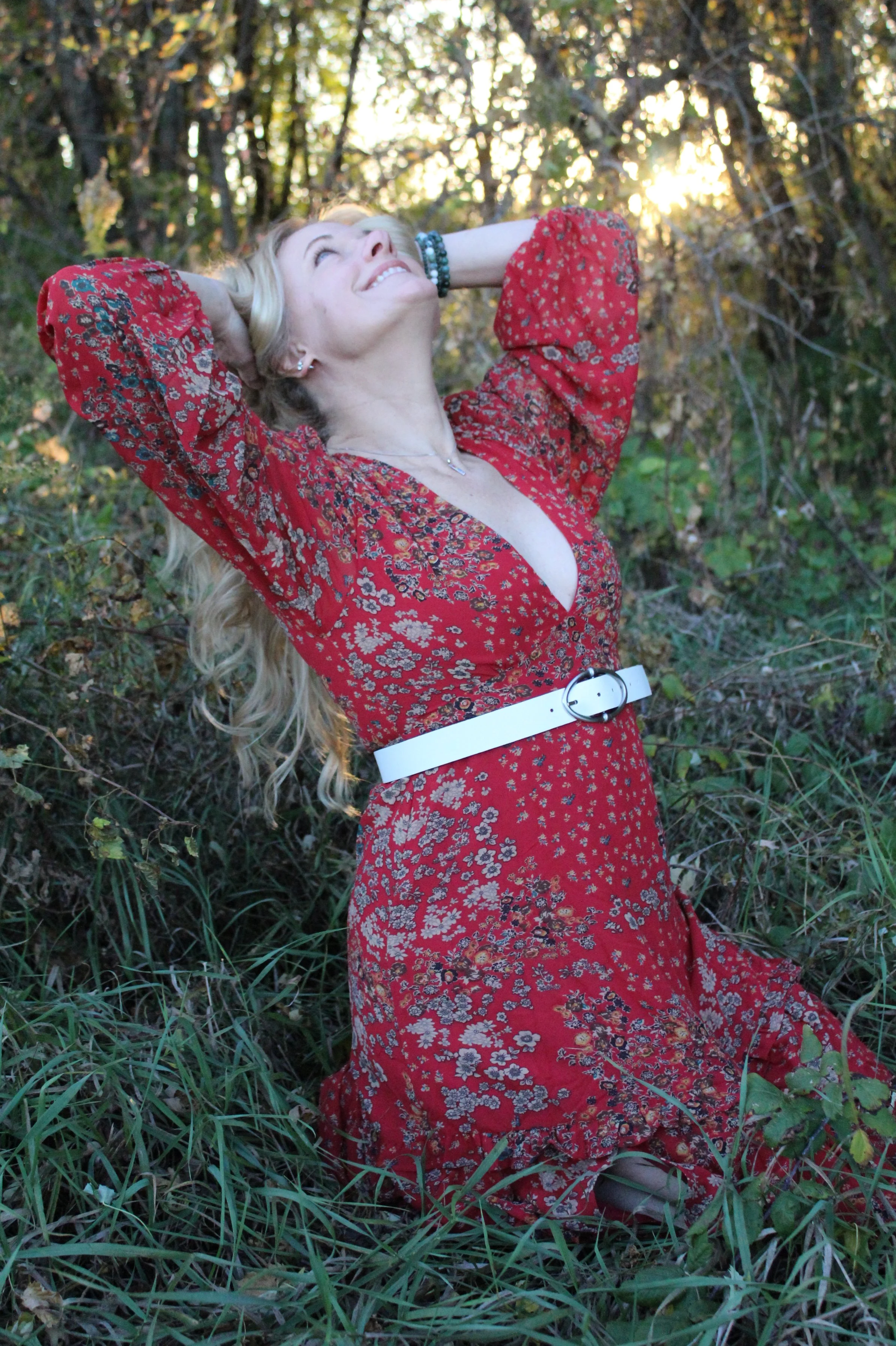A woman with long blonde hair, wearing a red floral dress and a white belt, kneeling on grass in a wooded area with sunlight filtering through trees, smiling with her eyes closed and arms behind her head.
