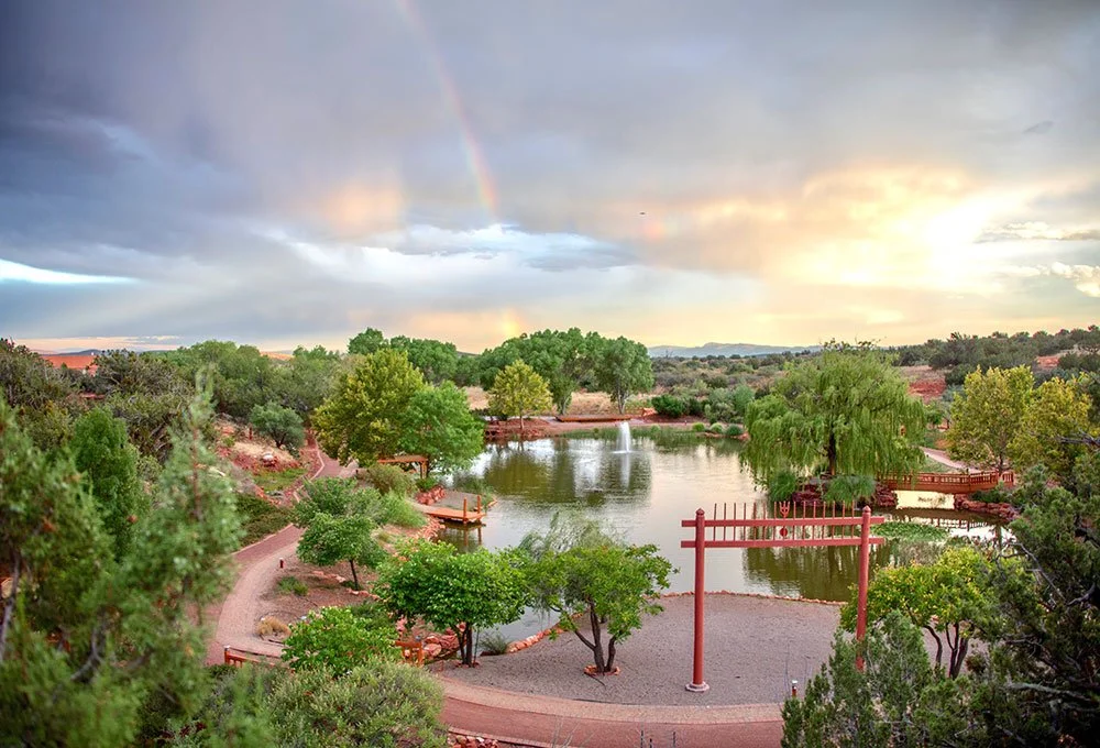 Sedona Mago retreat center with a pond, trees, walking paths, a red gate, and a rainbow in the sky during sunset.