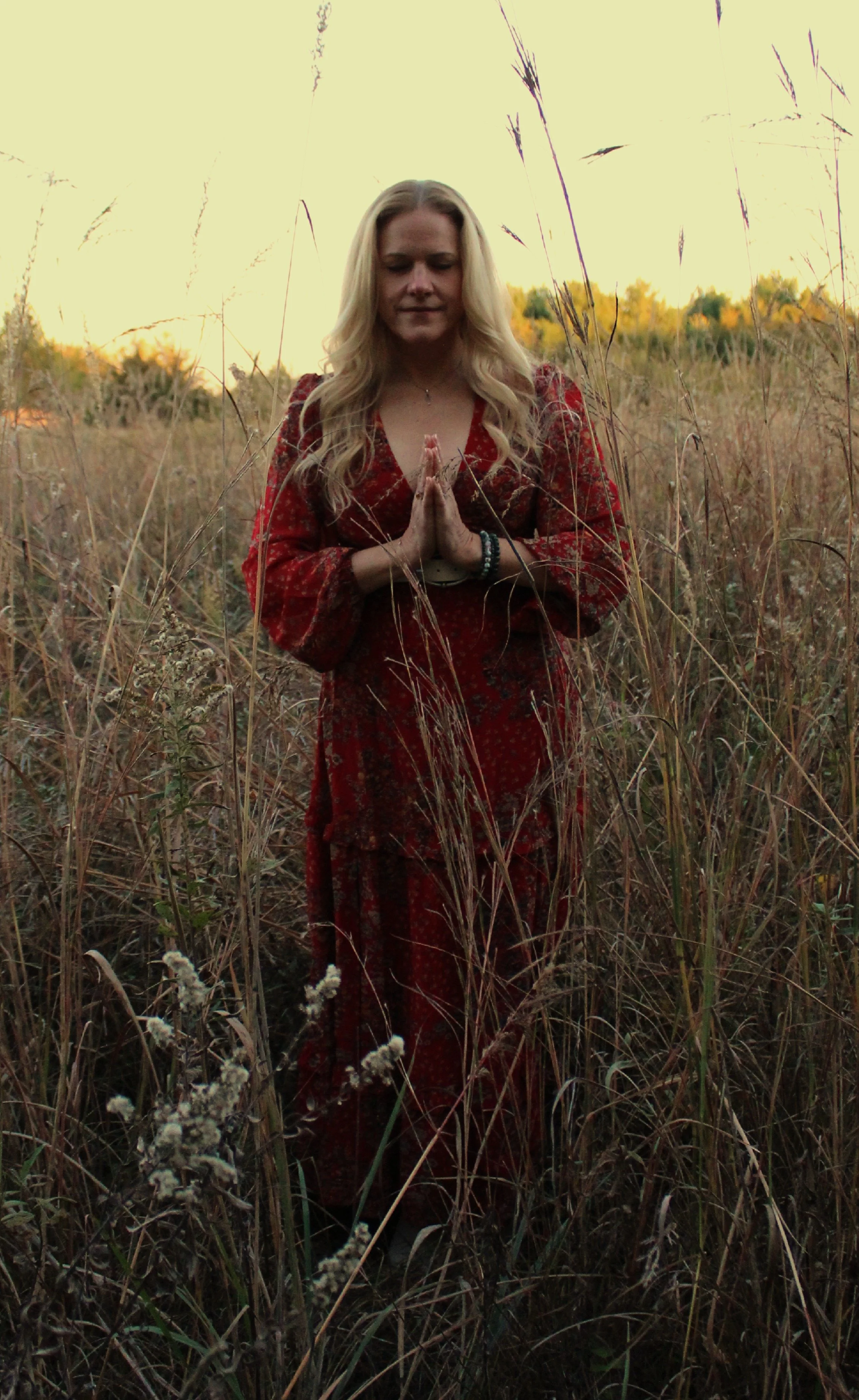 A woman with long blonde hair, wearing a red patterned dress, standing in a tall grass field at sunset with her hands in a prayer position.