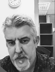 Black and white photo of a man with short, gray hair and a beard, looking directly at the camera, taken indoors with bookshelves and a clock in the background.