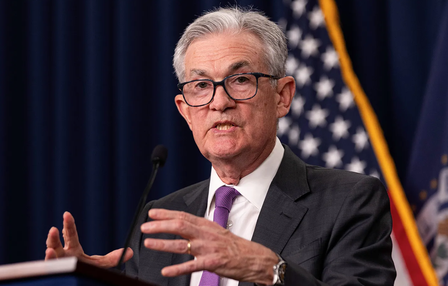 A senior man with gray hair, glasses, and a dark suit speaking at a podium with a U.S. flag in the background.