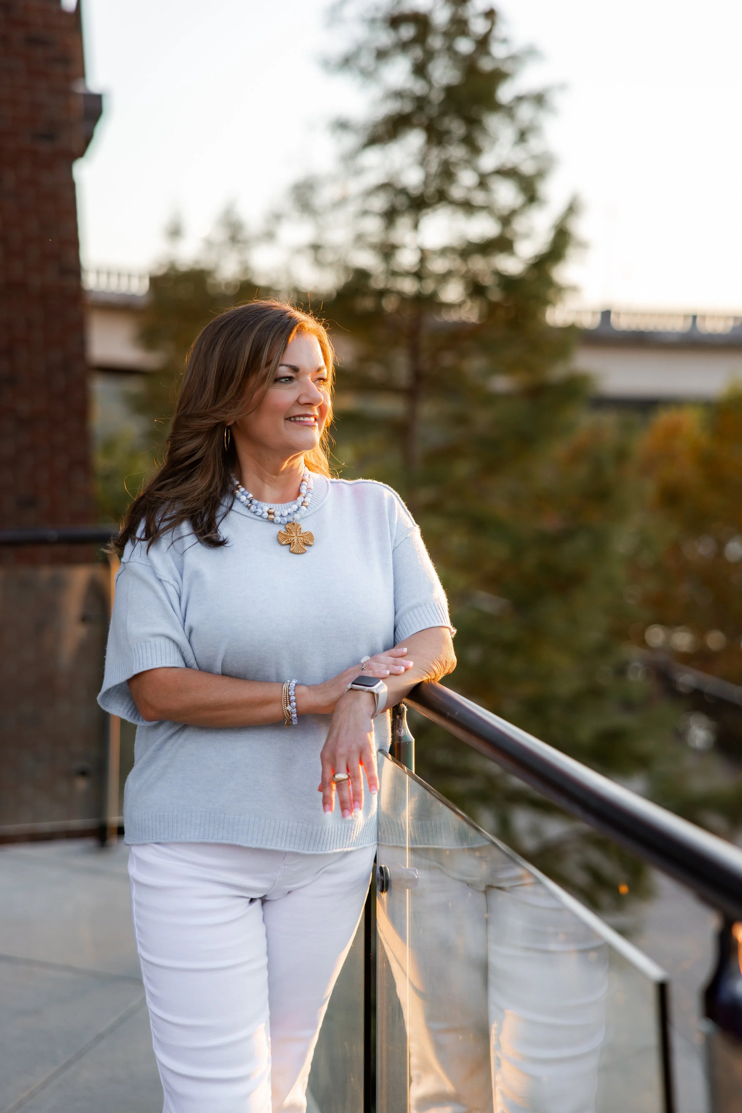 A woman with long brown hair, wearing a white top, white pants, and jewelry, stands outdoors on a balcony during sunset, looking to her right with a smile.