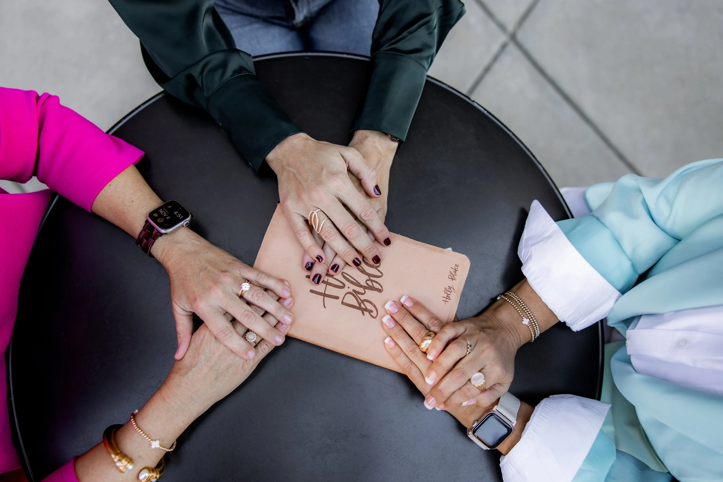 Multiple hands resting on a book titled "Holly Blake Big" on a round black table, with some wearing rings and bracelets, and a smartwatch visible.