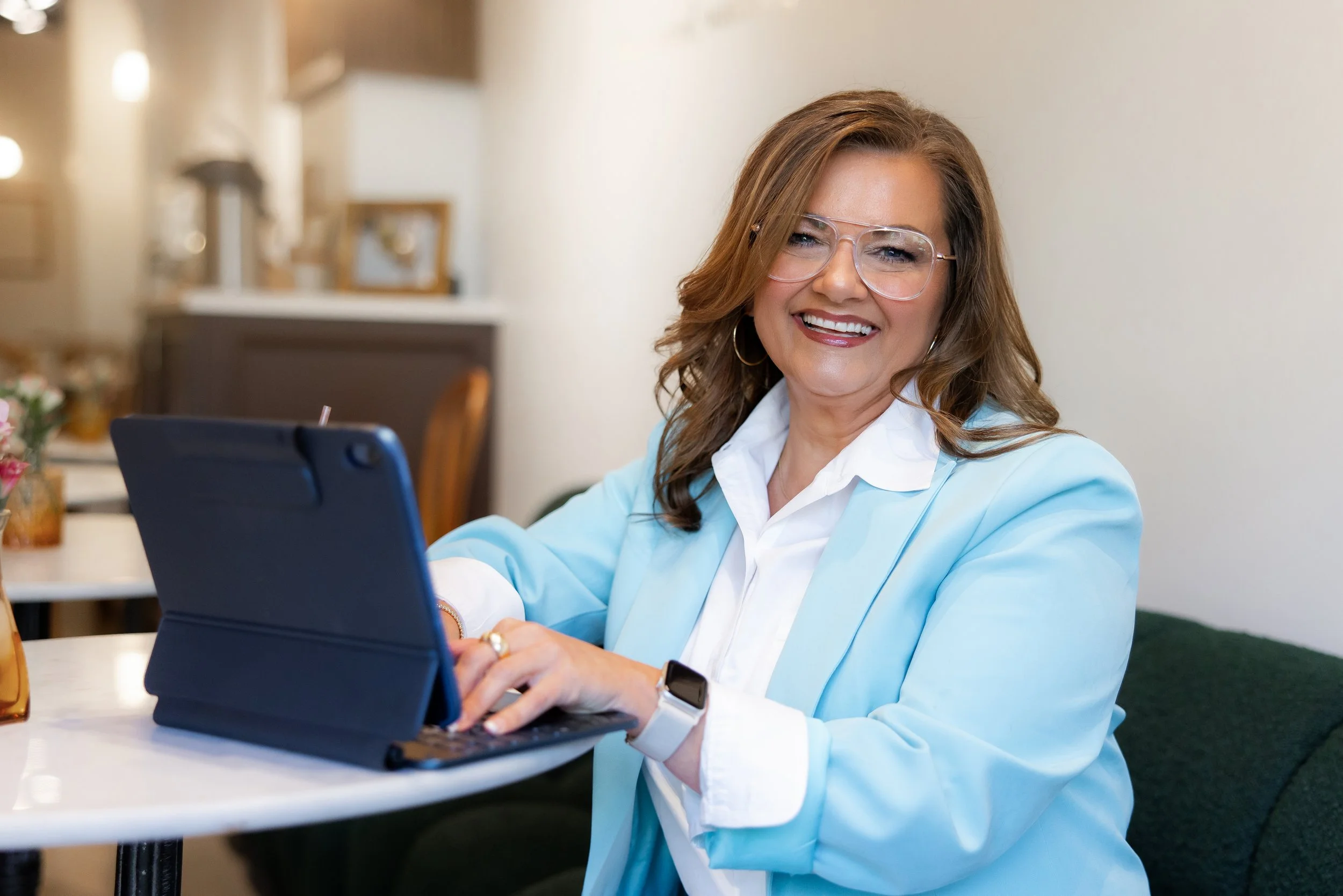 A smiling woman with glasses and a light blue blazer sitting at a table using a tablet in a bright indoor setting.