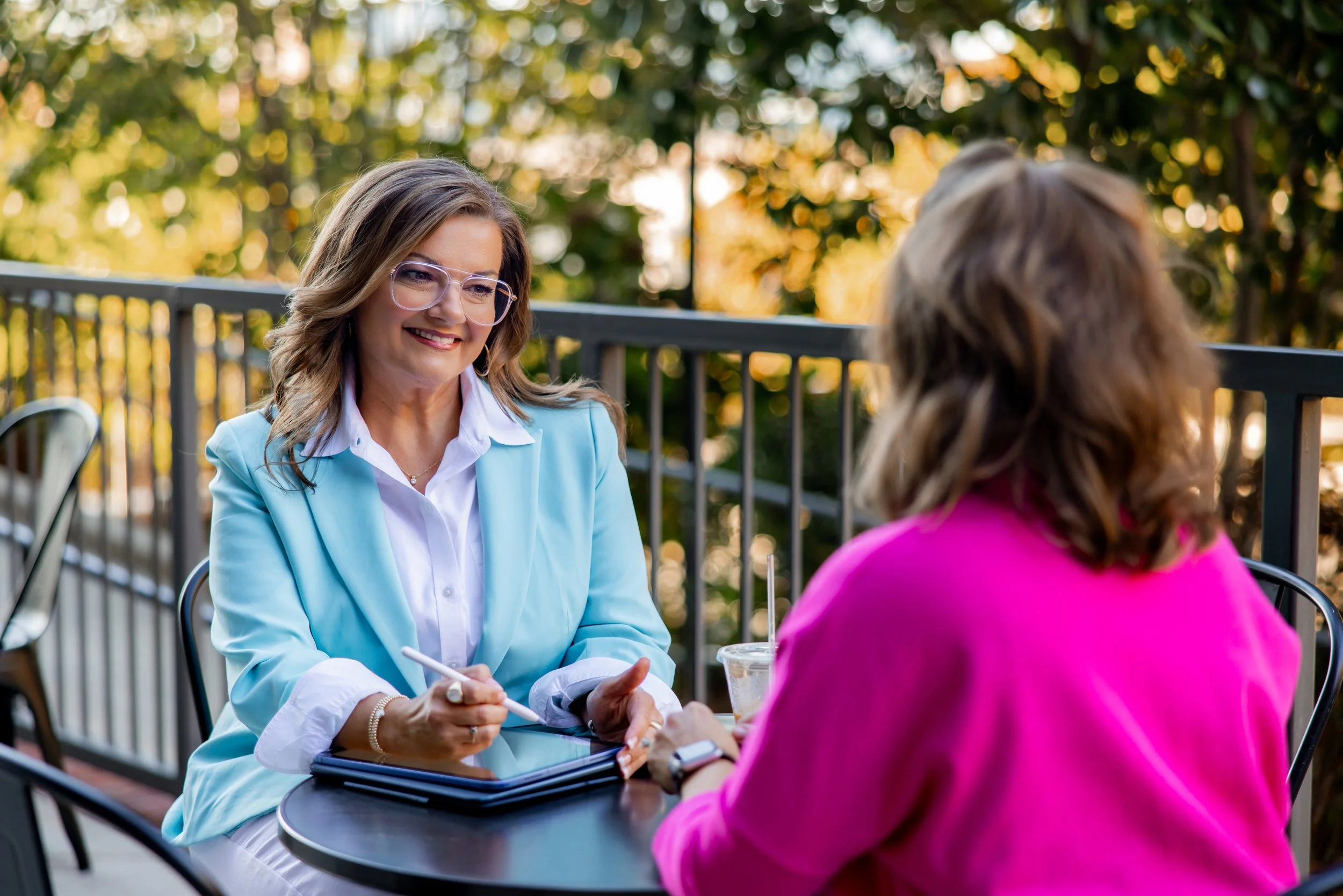 Two women sitting at an outdoor table, engaged in conversation, with a background of trees and warm sunlight.