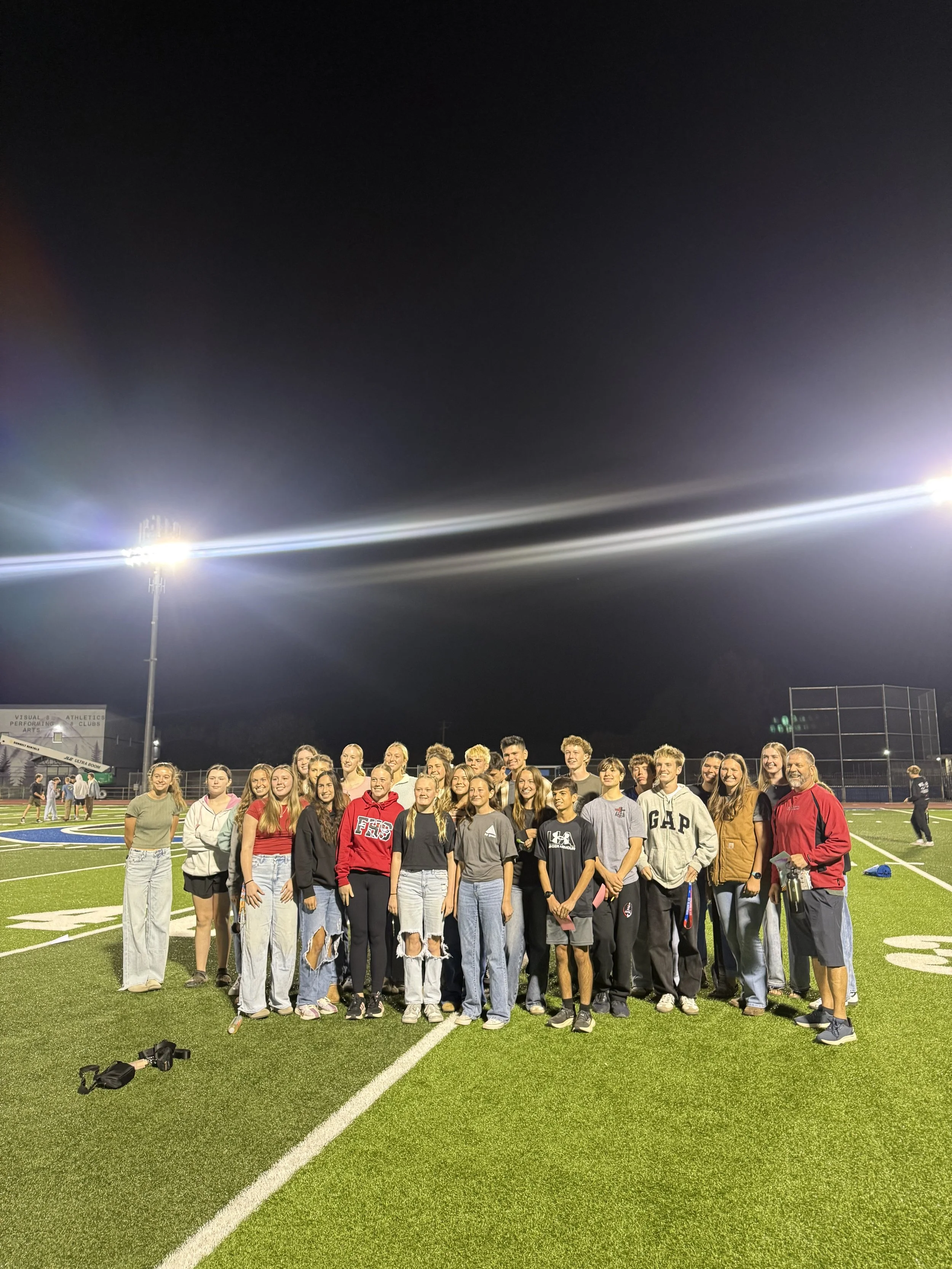 Group of teenagers and adults standing on a football field at night, under bright stadium lights, posing for a photo.