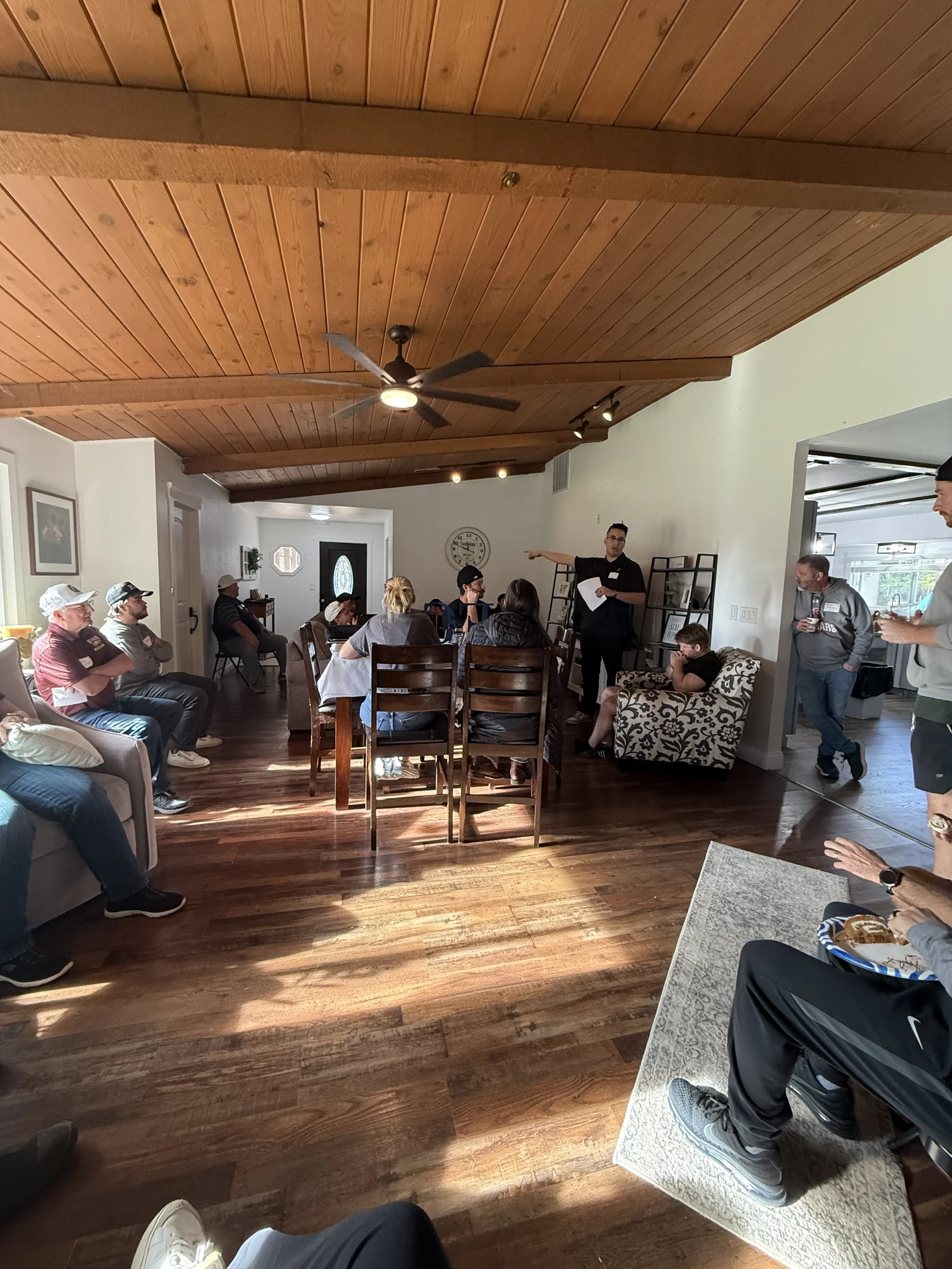 People sitting and listening to a speaker in a living room with a wooden ceiling, hardwood floor, and a mix of chairs and sofas, with a person pointing at the speaker.