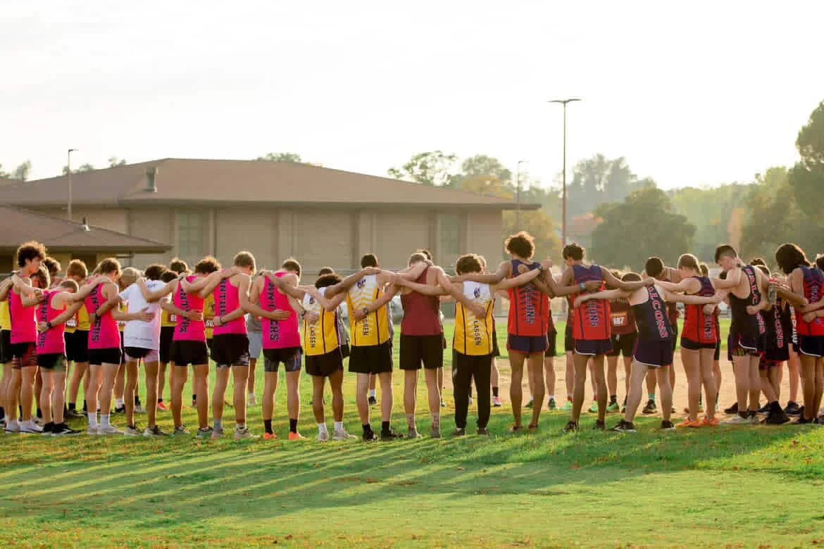 A group of athletes standing in a circle on a running track, with their arms around each other's shoulders, during what appears to be a team huddle or prayer.
