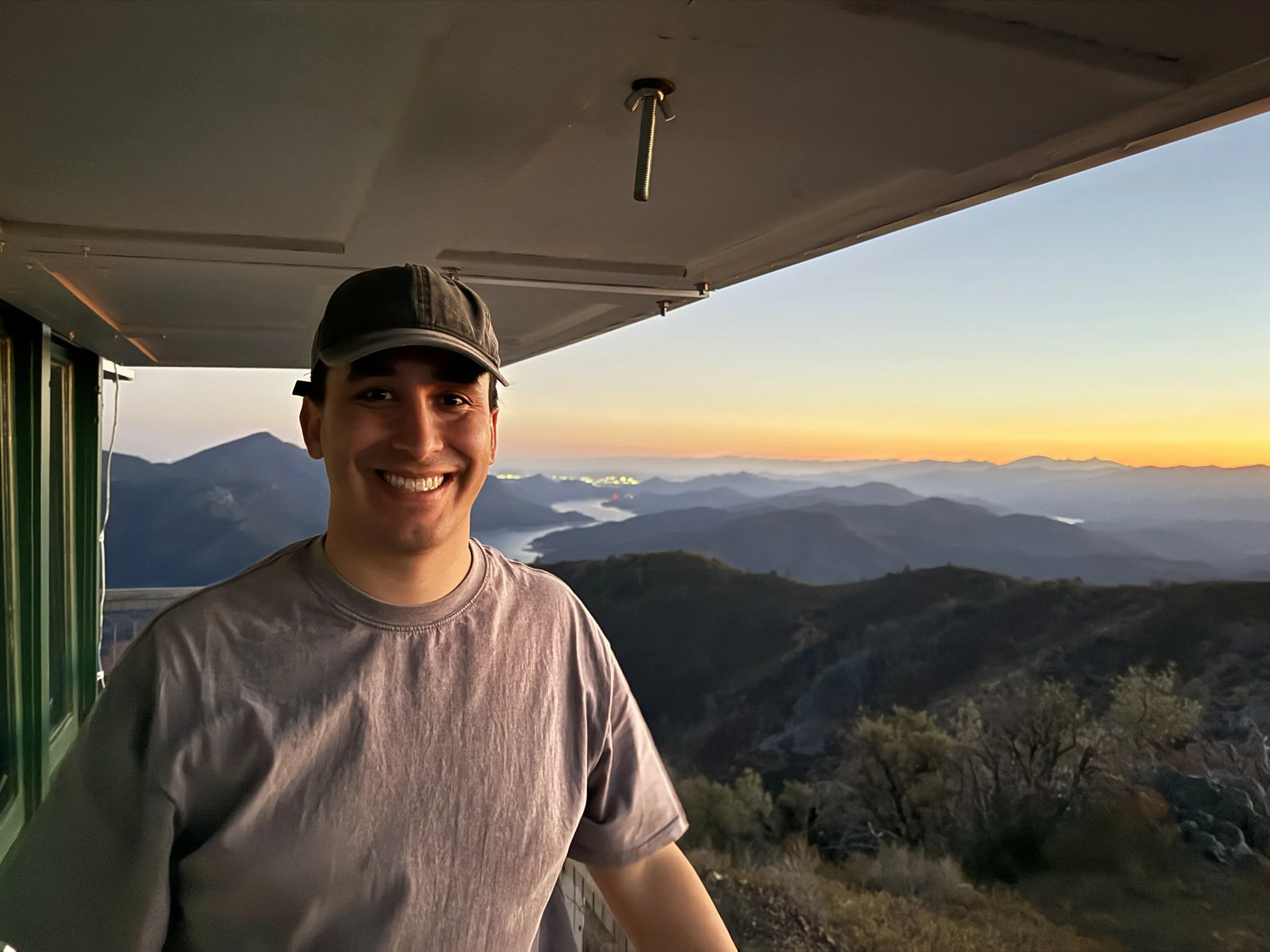 A smiling young man wearing a cap and t-shirt standing on a balcony with mountains and a colorful sunset in the background.