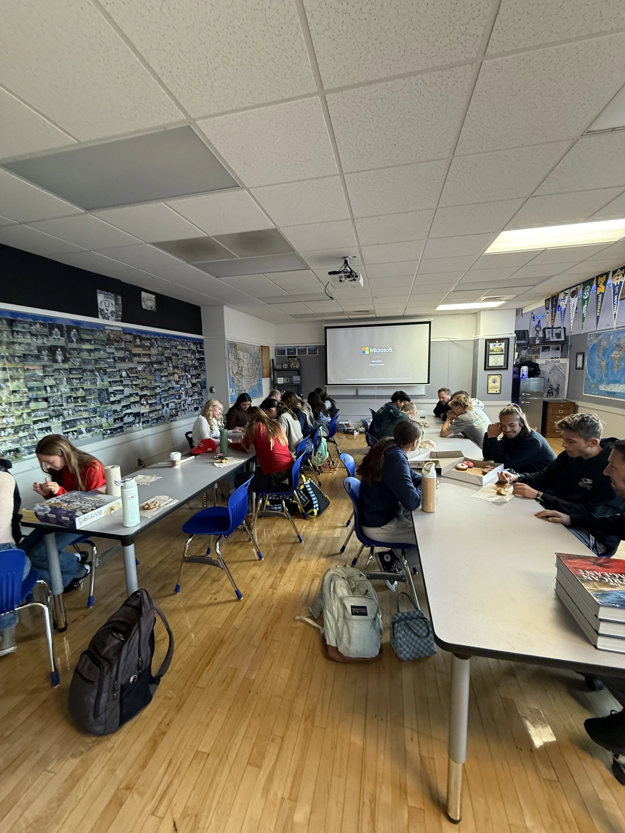A classroom filled with students sitting at tables, some eating snacks, with a projection screen displaying the Microsoft logo at the front of the room.