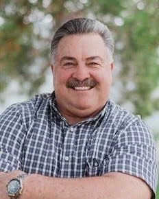 Smiling middle-aged man with gray hair and mustache, wearing a checkered shirt, standing outdoors with trees in the background.