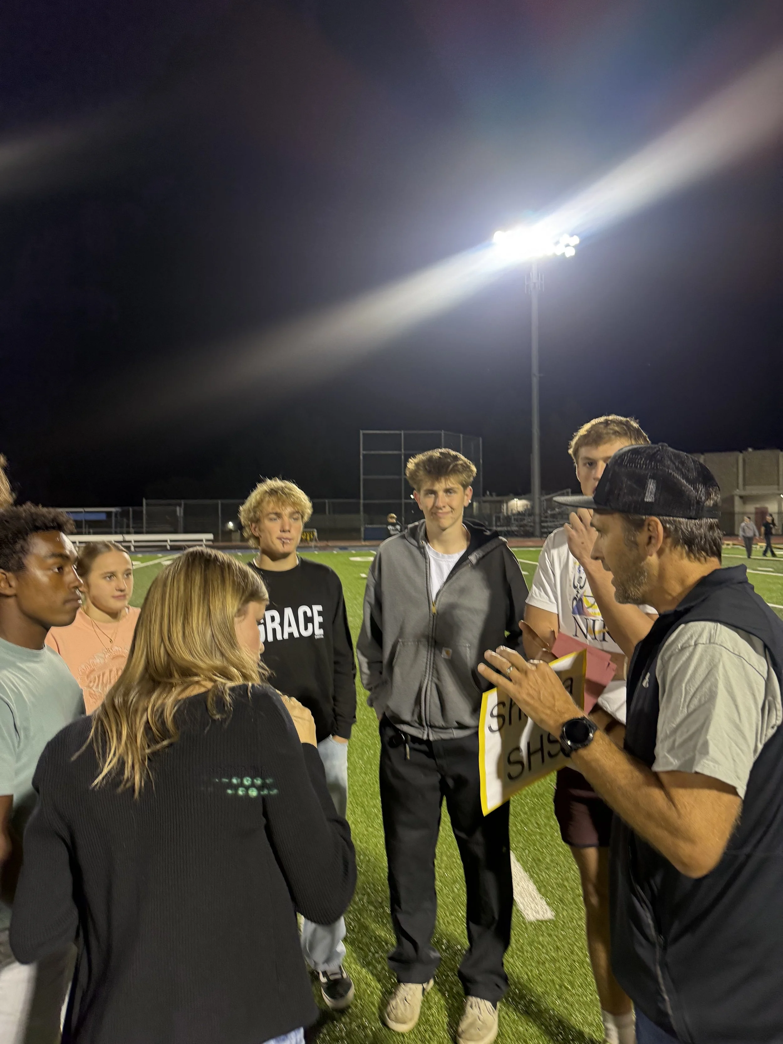 Group of young students listening to coach with a sign that reads 'SHS' on a football field at night, under bright stadium lights.