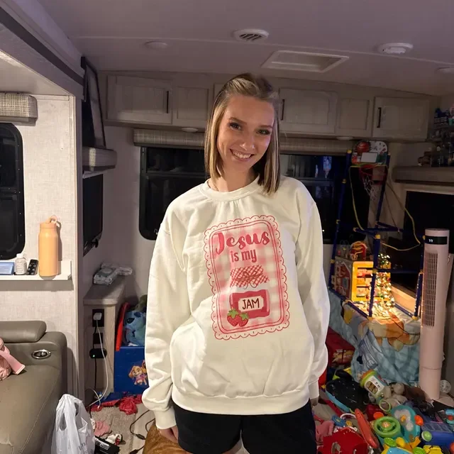 A young woman with shoulder-length blonde hair smiling in a camper or RV interior, wearing a white sweatshirt with red text and graphics reading "Jesus is my jam," and black shorts, surrounded by clutter including toys, a water bottle, and household items.