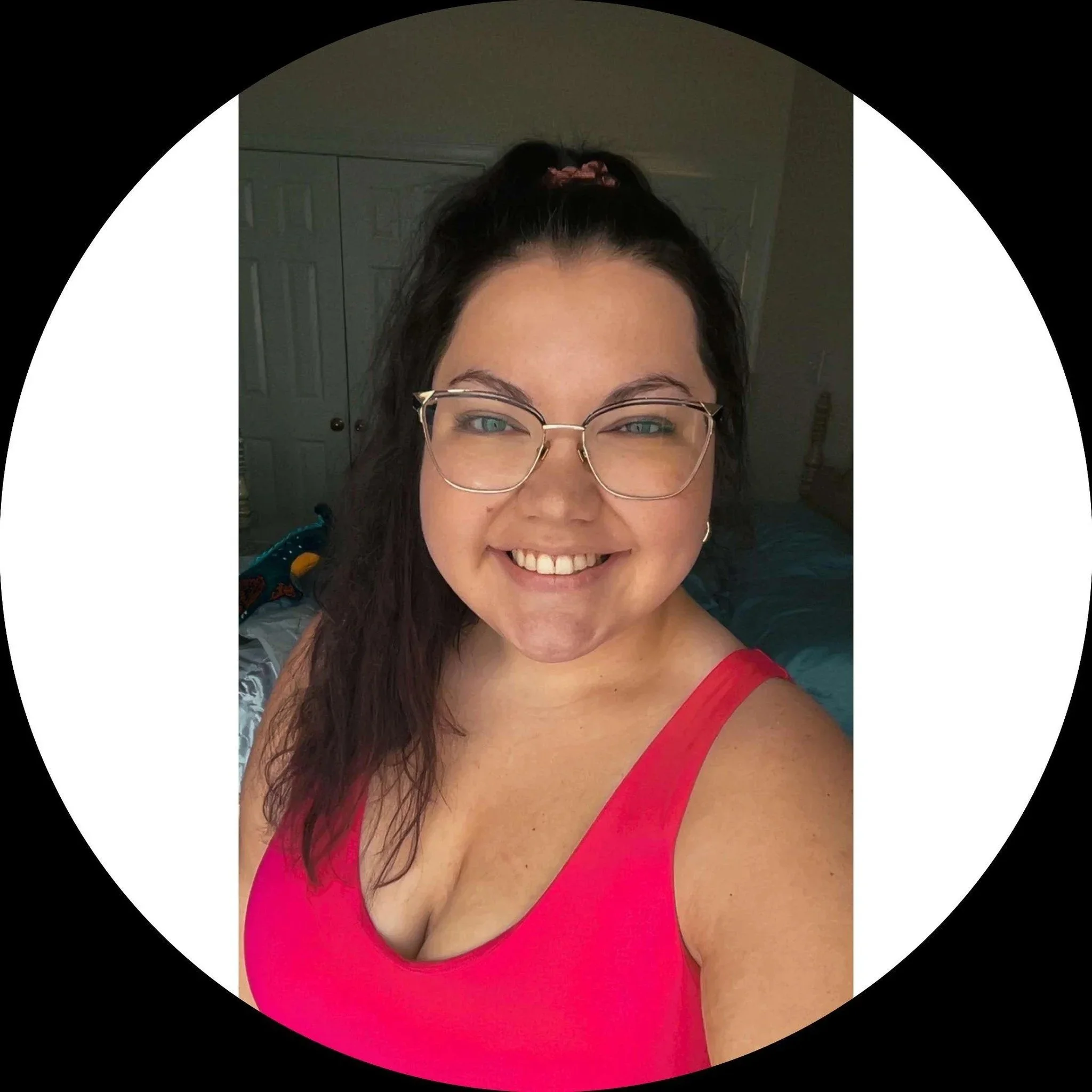 A woman with glasses and dark, wavy hair smiling, wearing a pink sleeveless top, inside a bedroom with closed doors in the background.