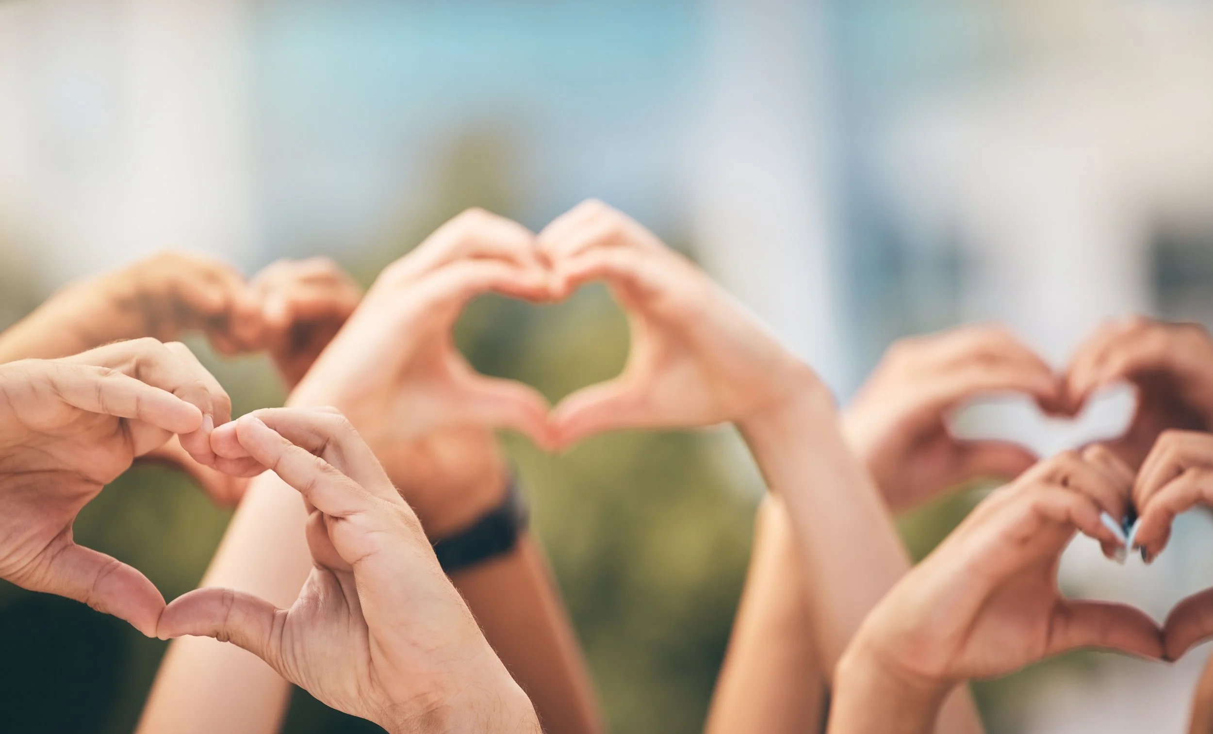 Multiple hands forming heart shapes in front of a blurred outdoor background.