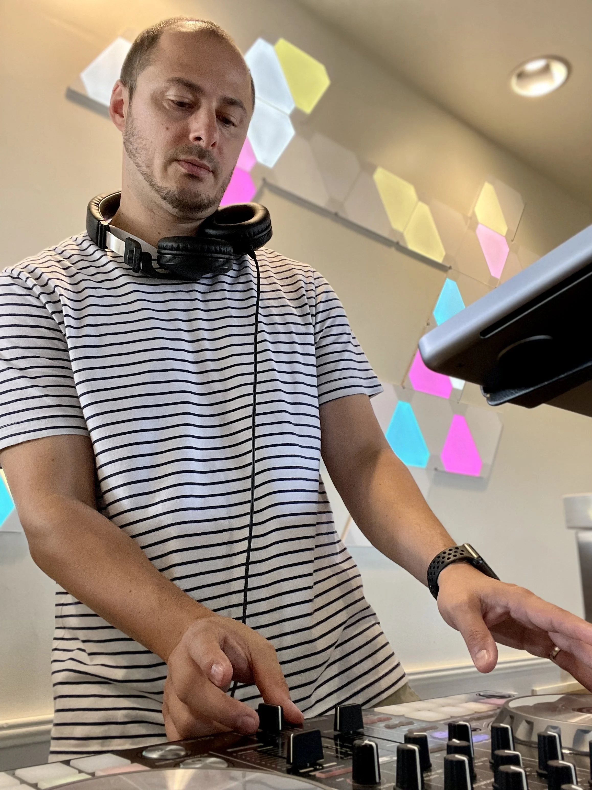 DJ MV with headphones around his neck, wearing a striped t-shirt, is DJing with a mixer and laptop in front of him, in a room with colorful hexagonal wall decorations.